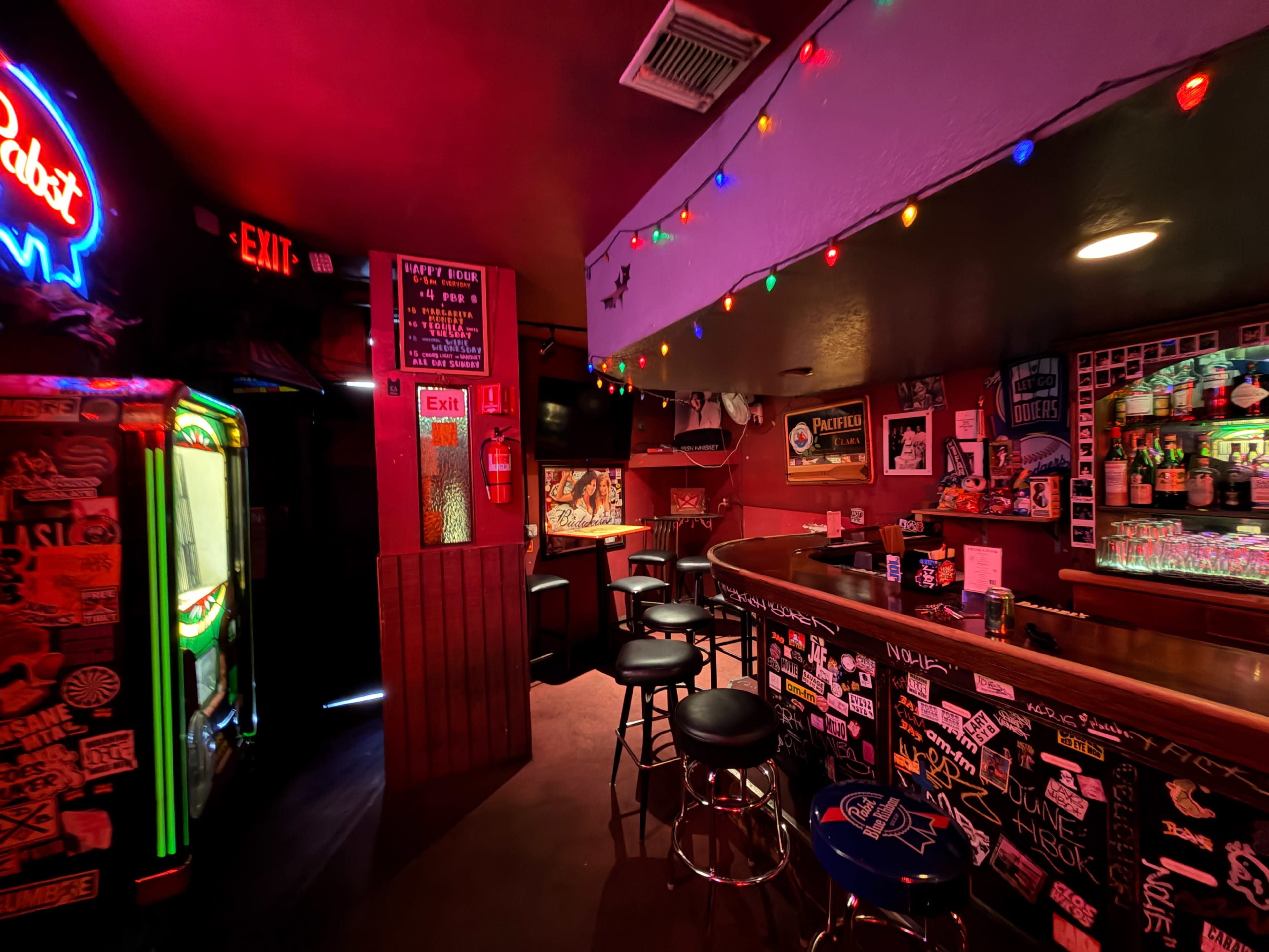 The image shows a dimly lit bar featuring a curved counter, bar stools, a jukebox, and walls adorned with various stickers and posters.
