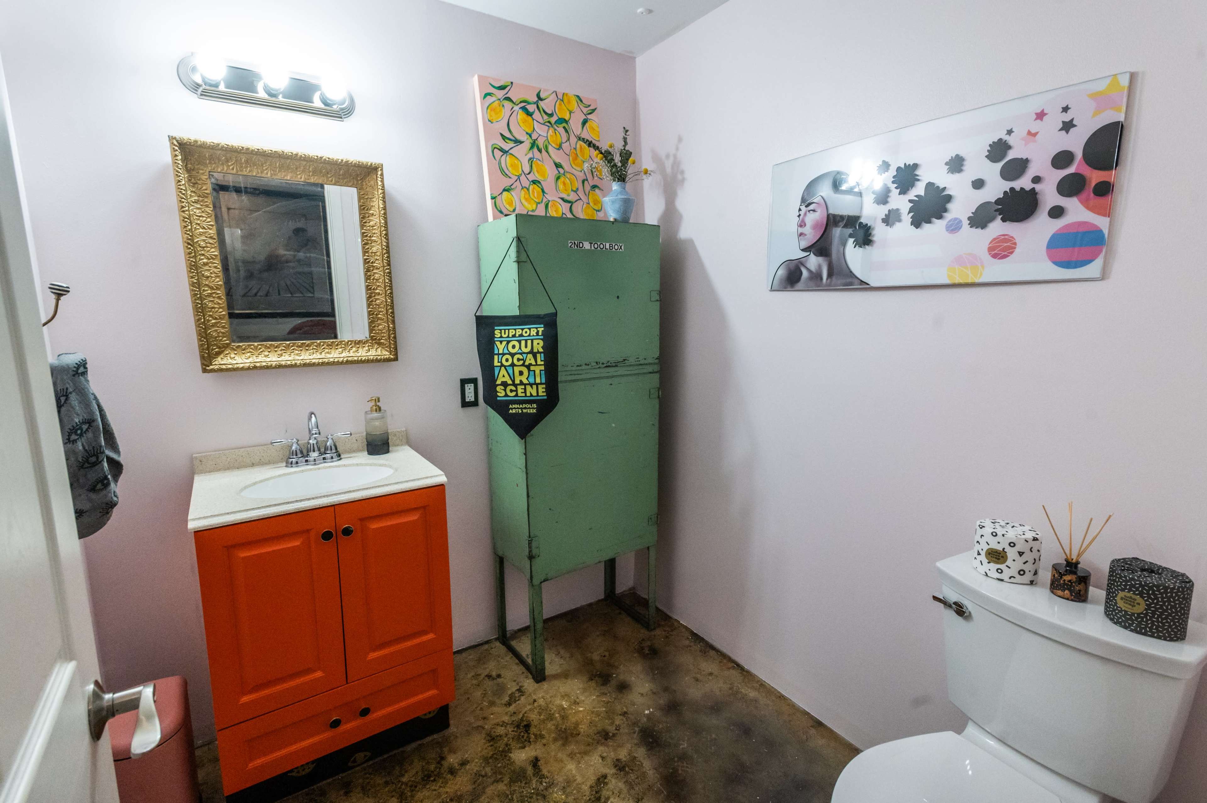 A small bathroom featuring a green metal cabinet, an orange vanity with a sink, and various artwork on the walls.