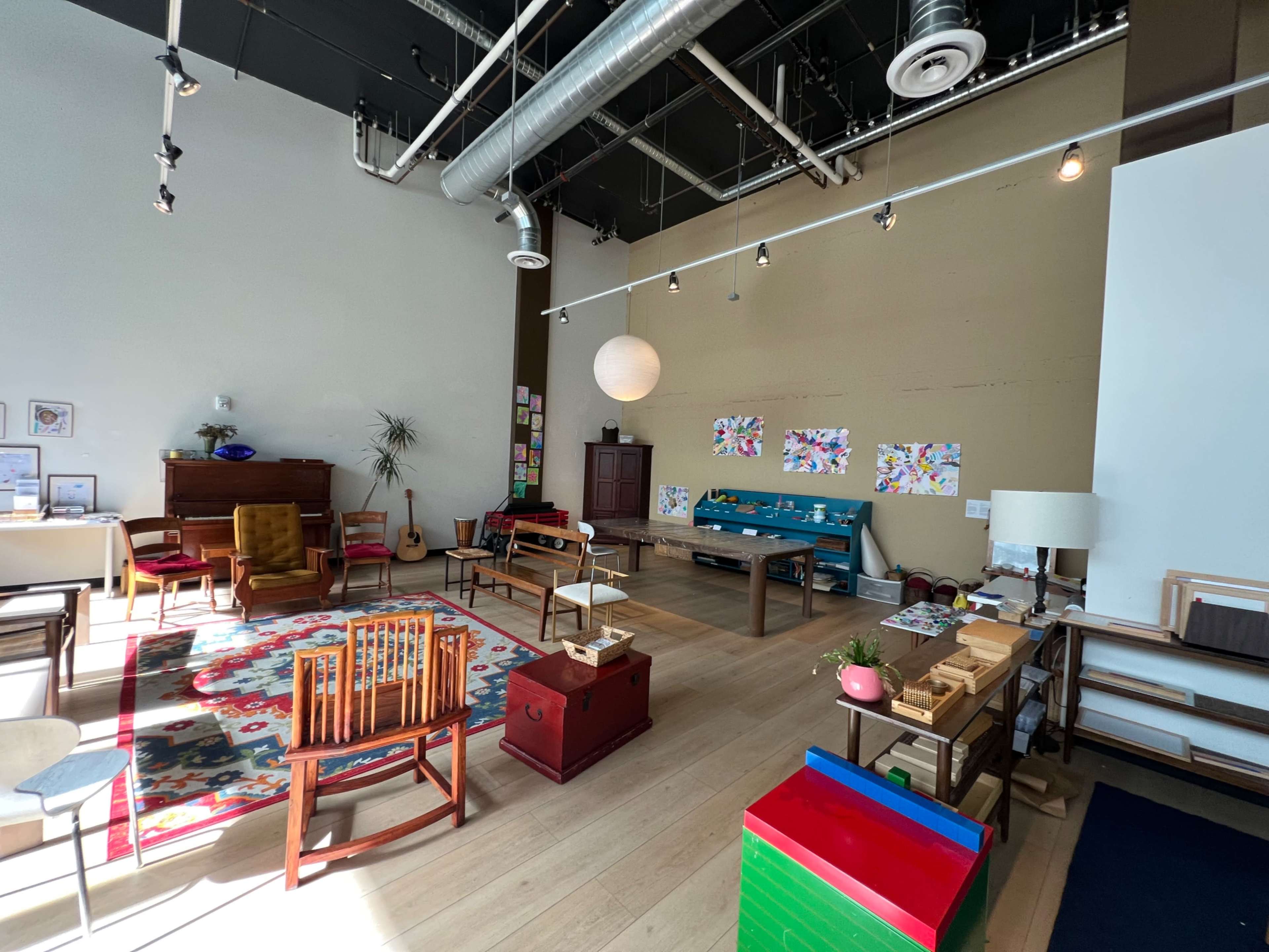 The image shows a spacious room with various pieces of furniture, including colorful chairs, a rug, and tables, alongside artwork on the walls and natural light streaming through large windows.