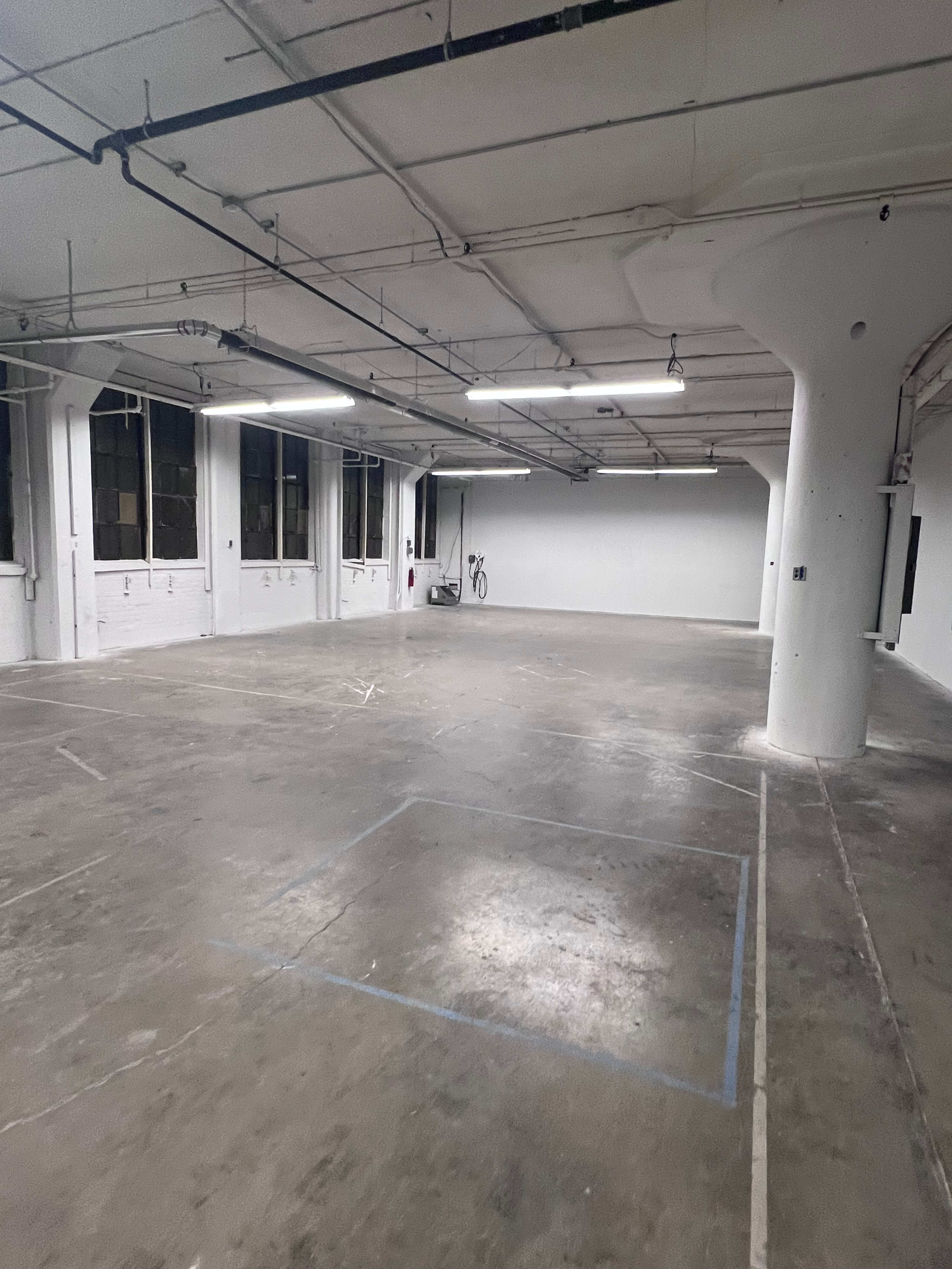 The image shows a spacious, empty industrial room with concrete flooring and exposed pipes, illuminated by overhead fluorescent lights.