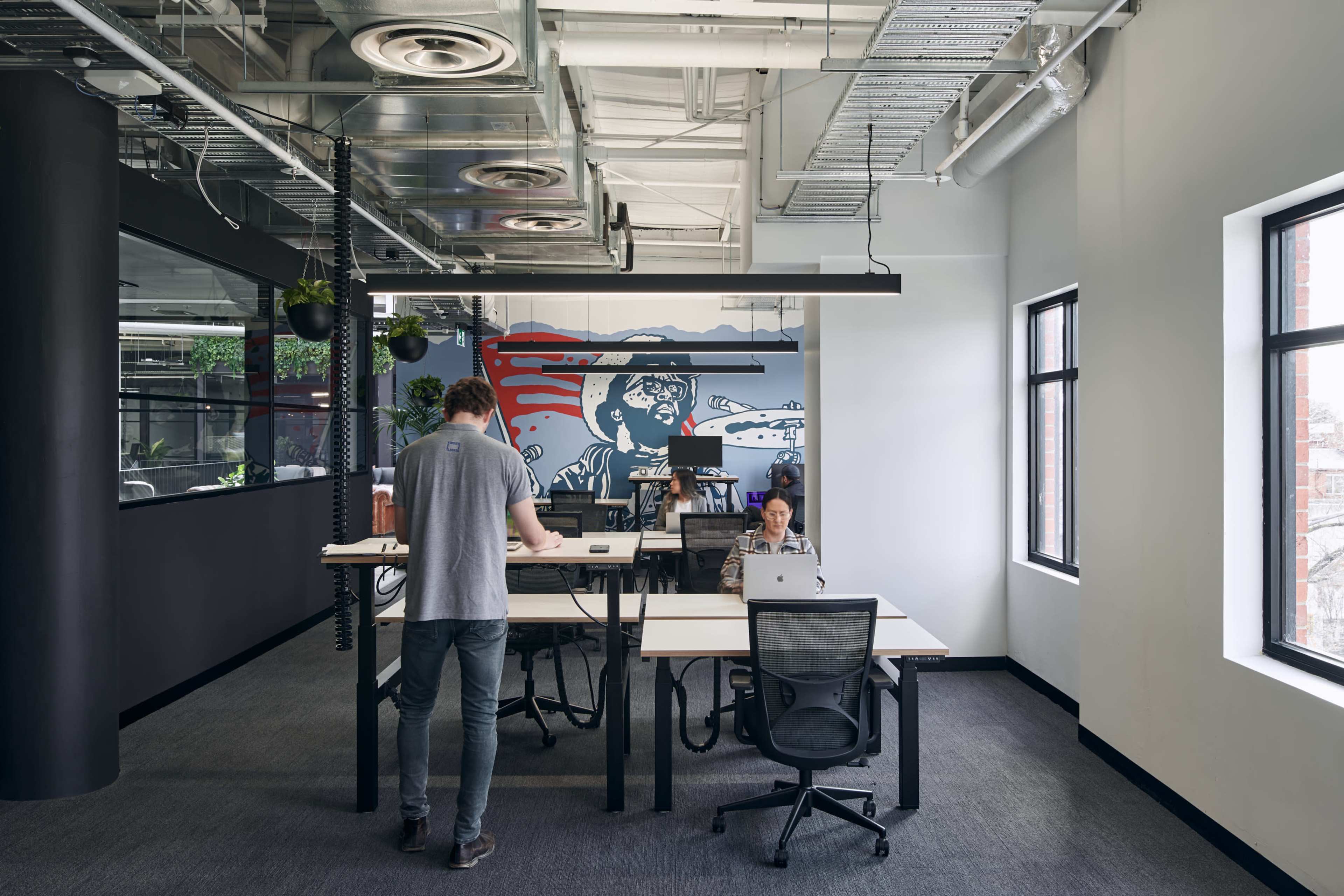 The image shows an open office space with two people working at desks and a mural on the wall in the background.