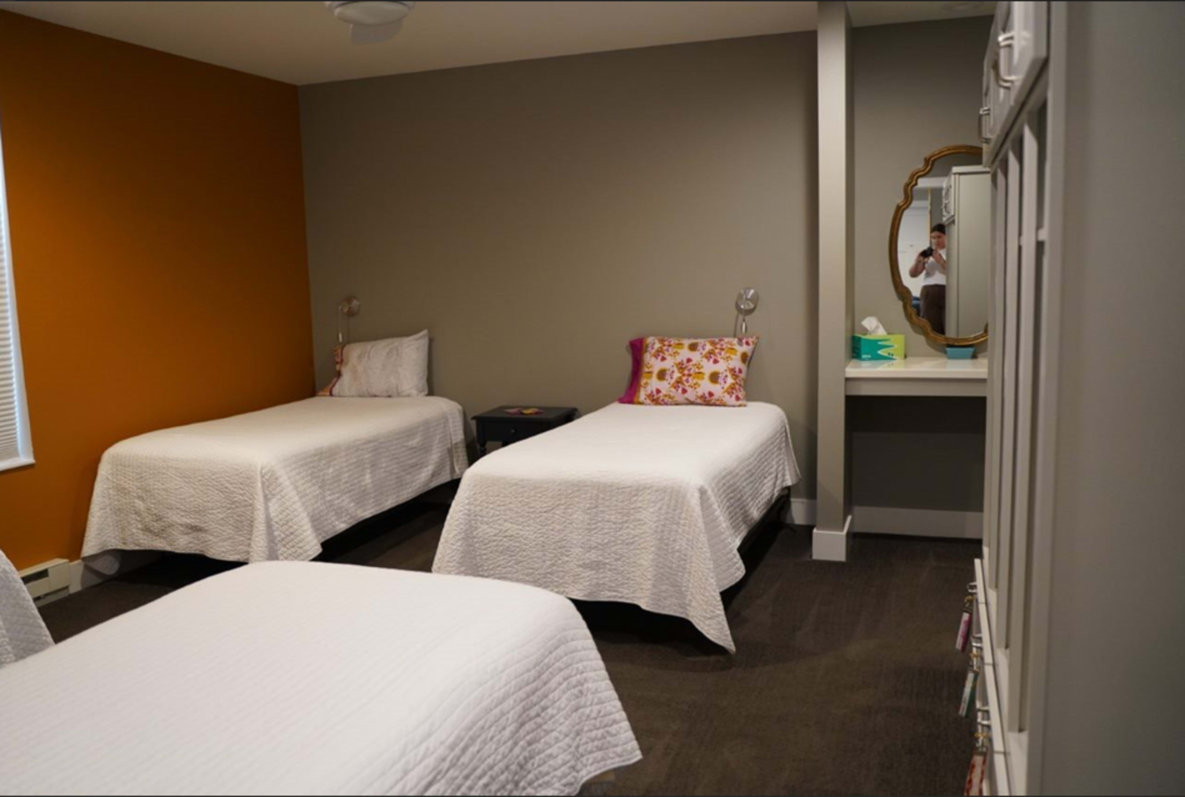 The image shows a room with three single beds arranged with white bedspreads, a small nightstand beside one bed, and a vanity with a mirror against a gray wall.