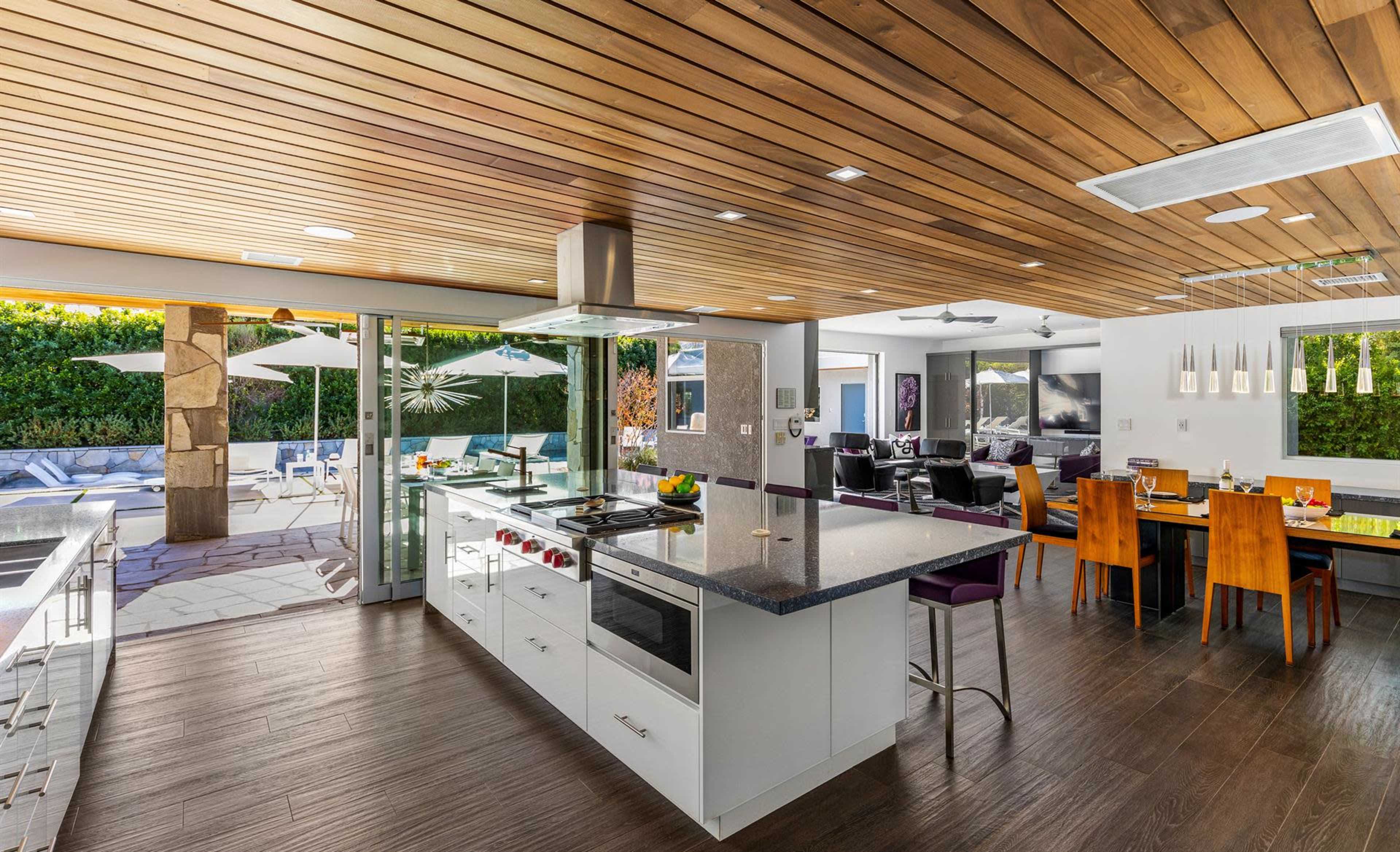 A modern kitchen with a large island and open layout connects to a living area and outdoor pool space through glass doors.