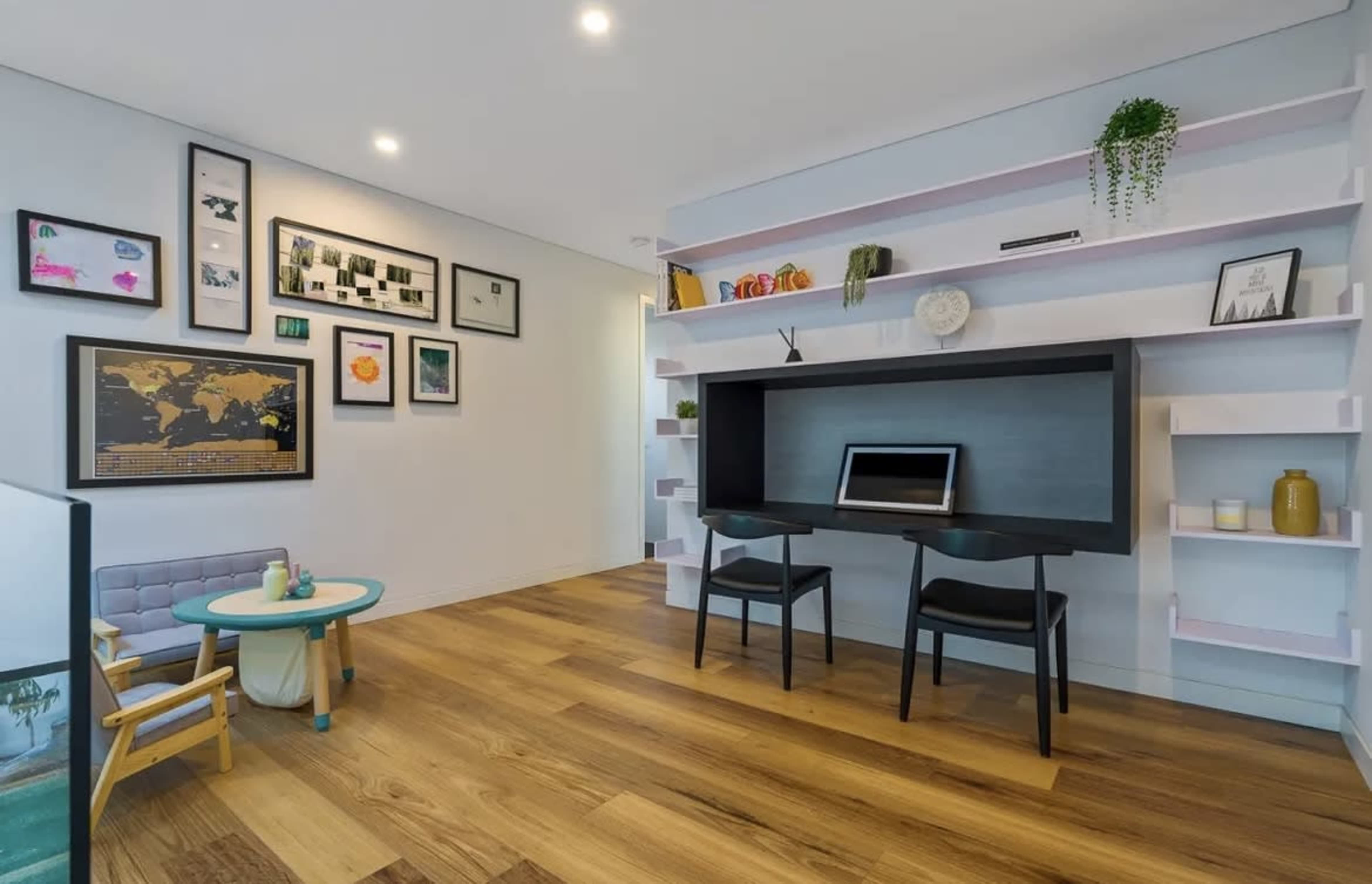 A modern home office features a built-in desk with two chairs, surrounded by framed art and decorative shelves on the walls.