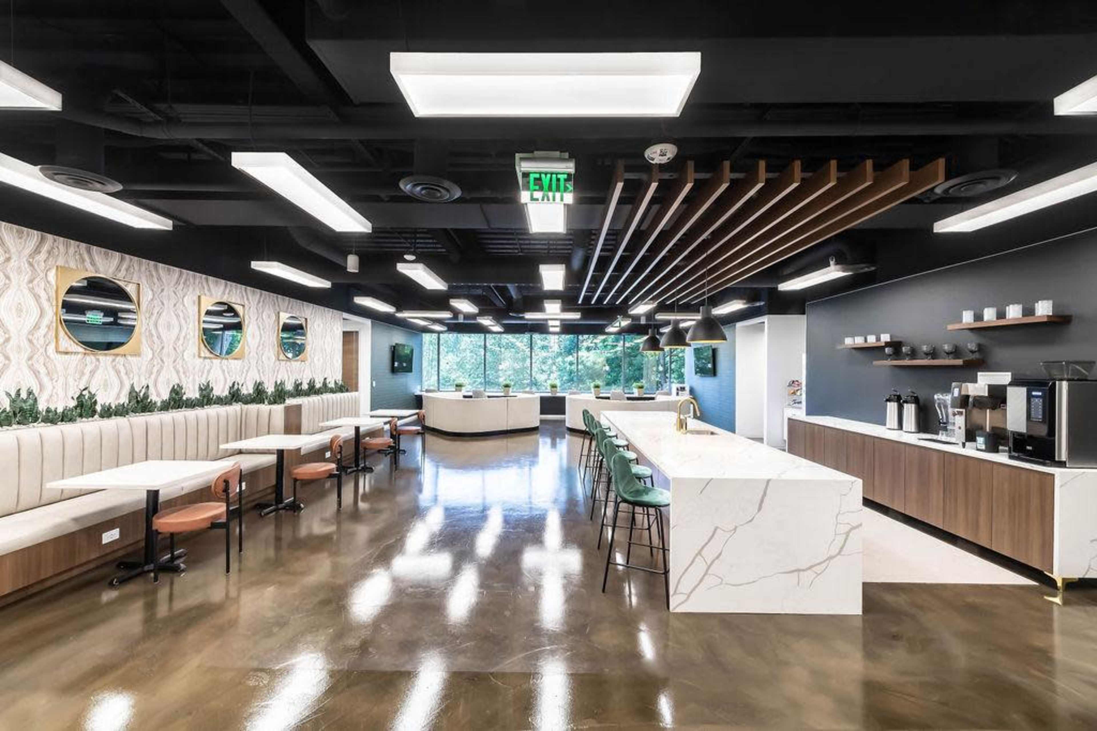 The image shows a modern office kitchen area with a long marble countertop, seating booths, and large windows letting in natural light.