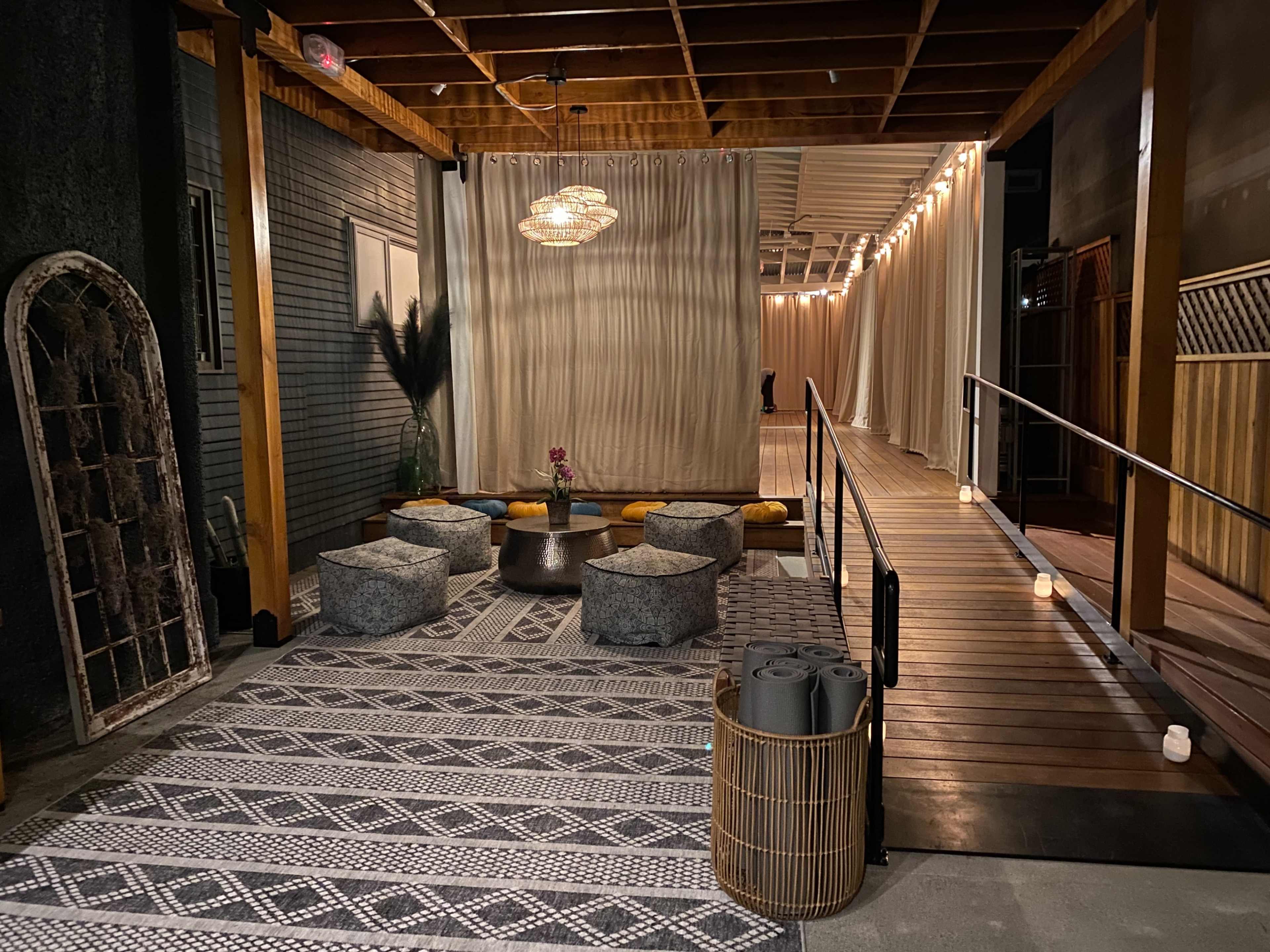 The image depicts a modern lounge area with a patterned rug, circular seating, and a pathway leading to a softly lit space adorned with light fixtures and decorative curtains.