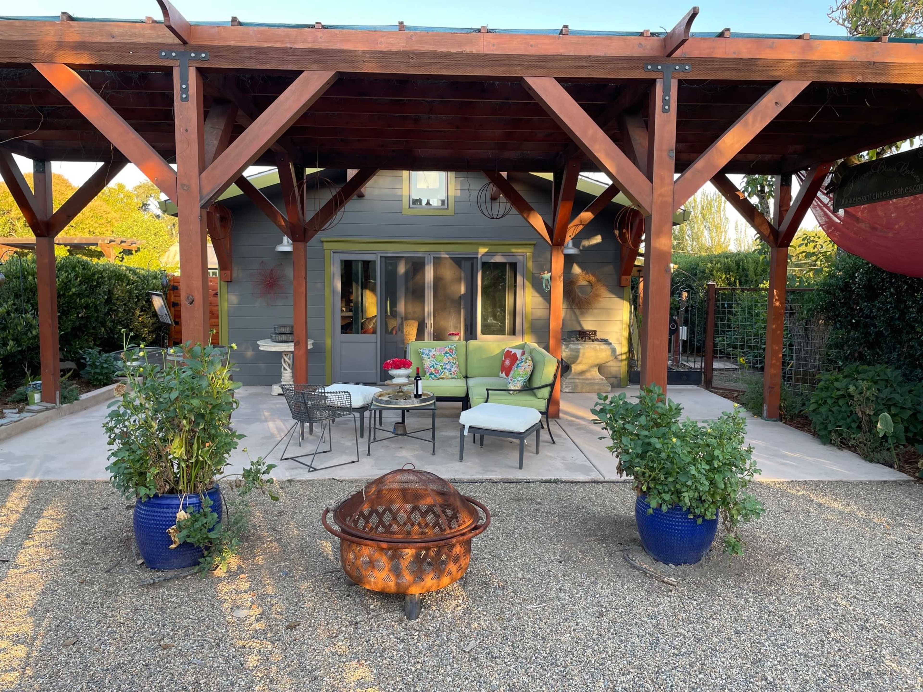 A patio area features a seating arrangement with green cushions, surrounded by potted plants and a fire pit, under a wooden pergola.