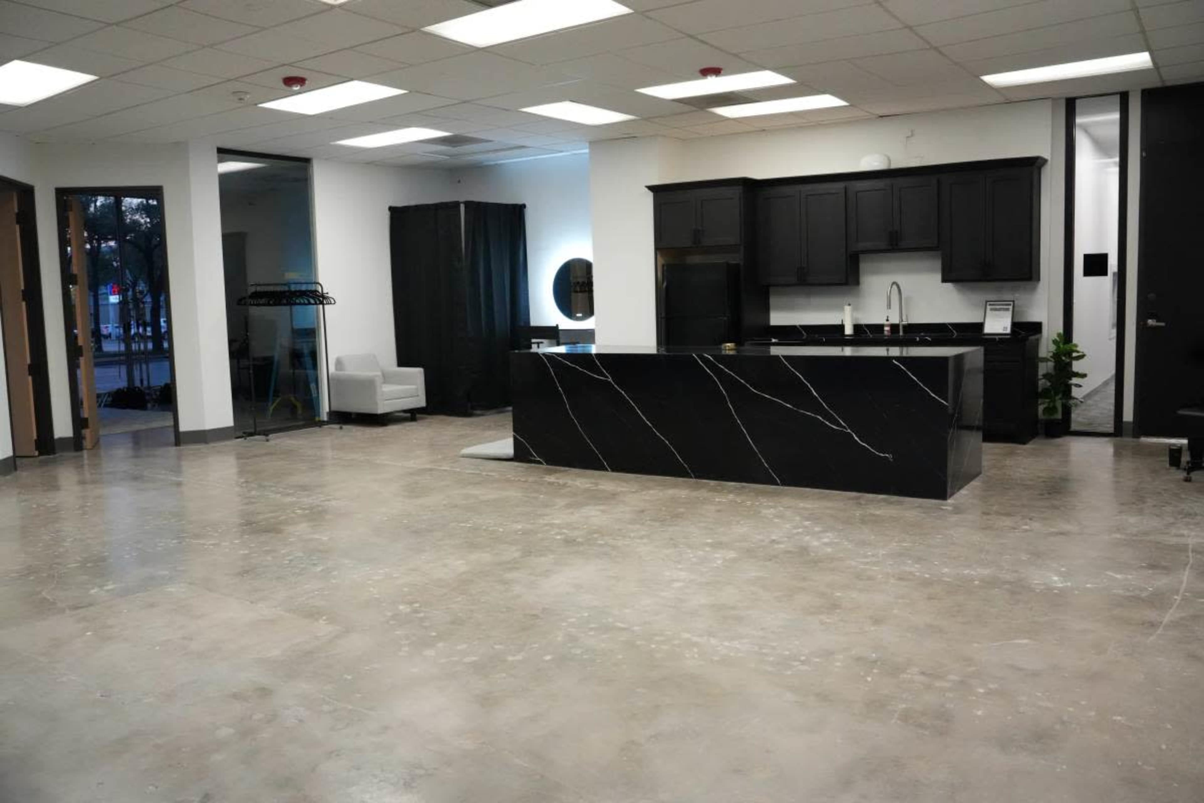 The image shows a spacious, minimalist interior with polished concrete flooring, featuring a black marble kitchen island and sparse furniture, including a white chair.