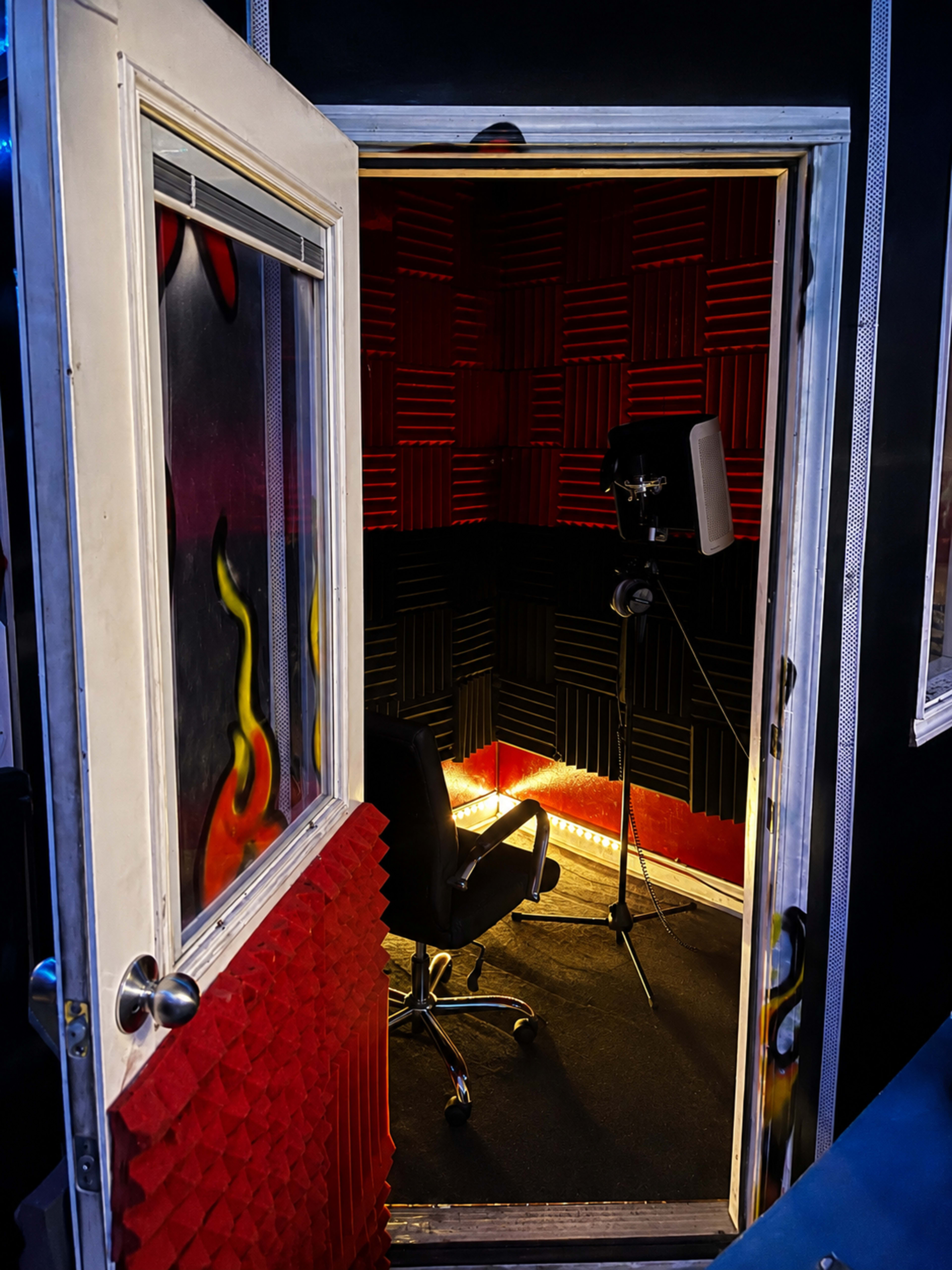 The image shows an open door leading into a soundproof recording studio with red and black acoustic foam panels and a microphone on a stand facing a black chair.