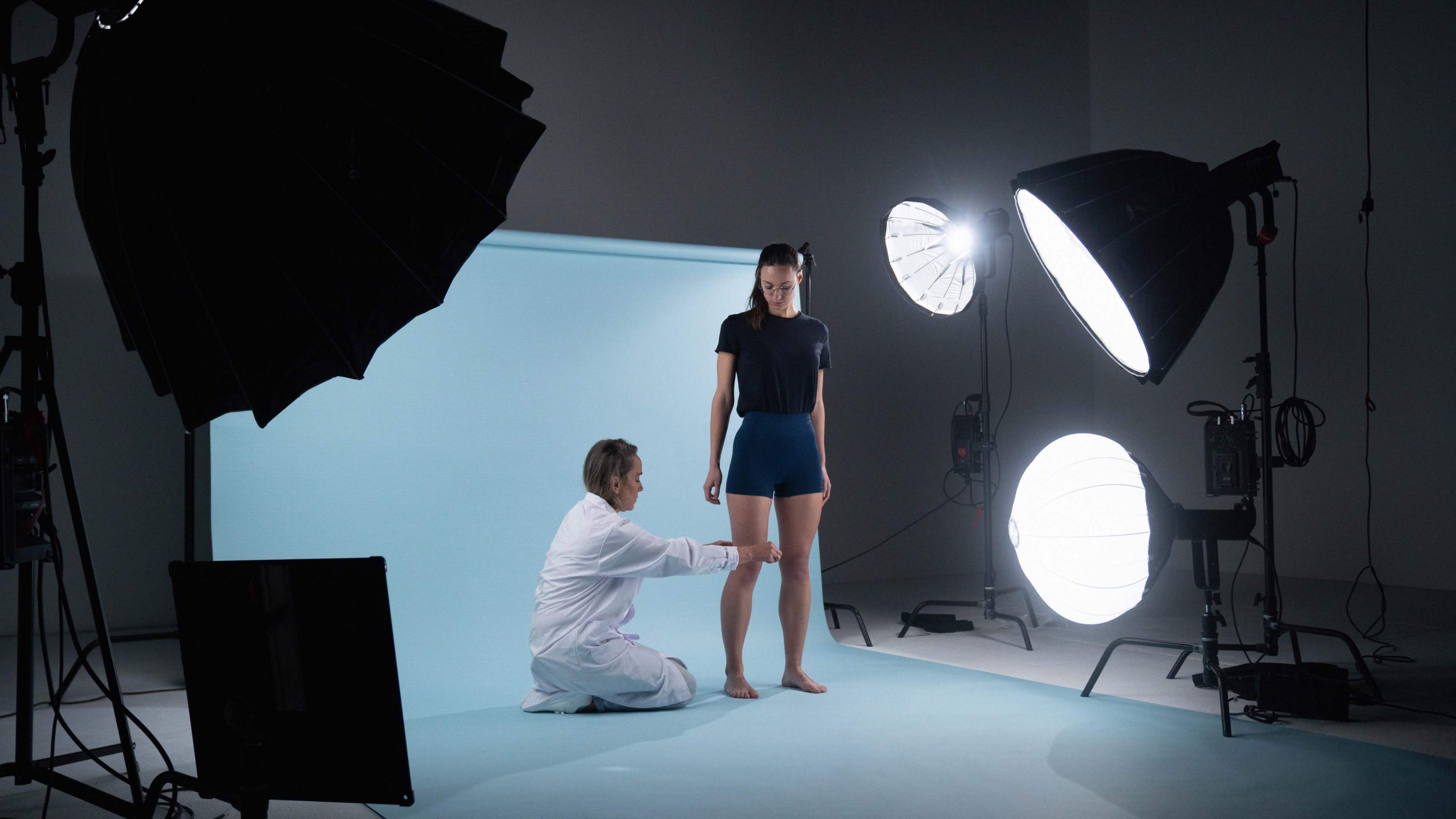 A photographer adjusts a model's pose in a studio setting with large softbox lights and a blue backdrop.