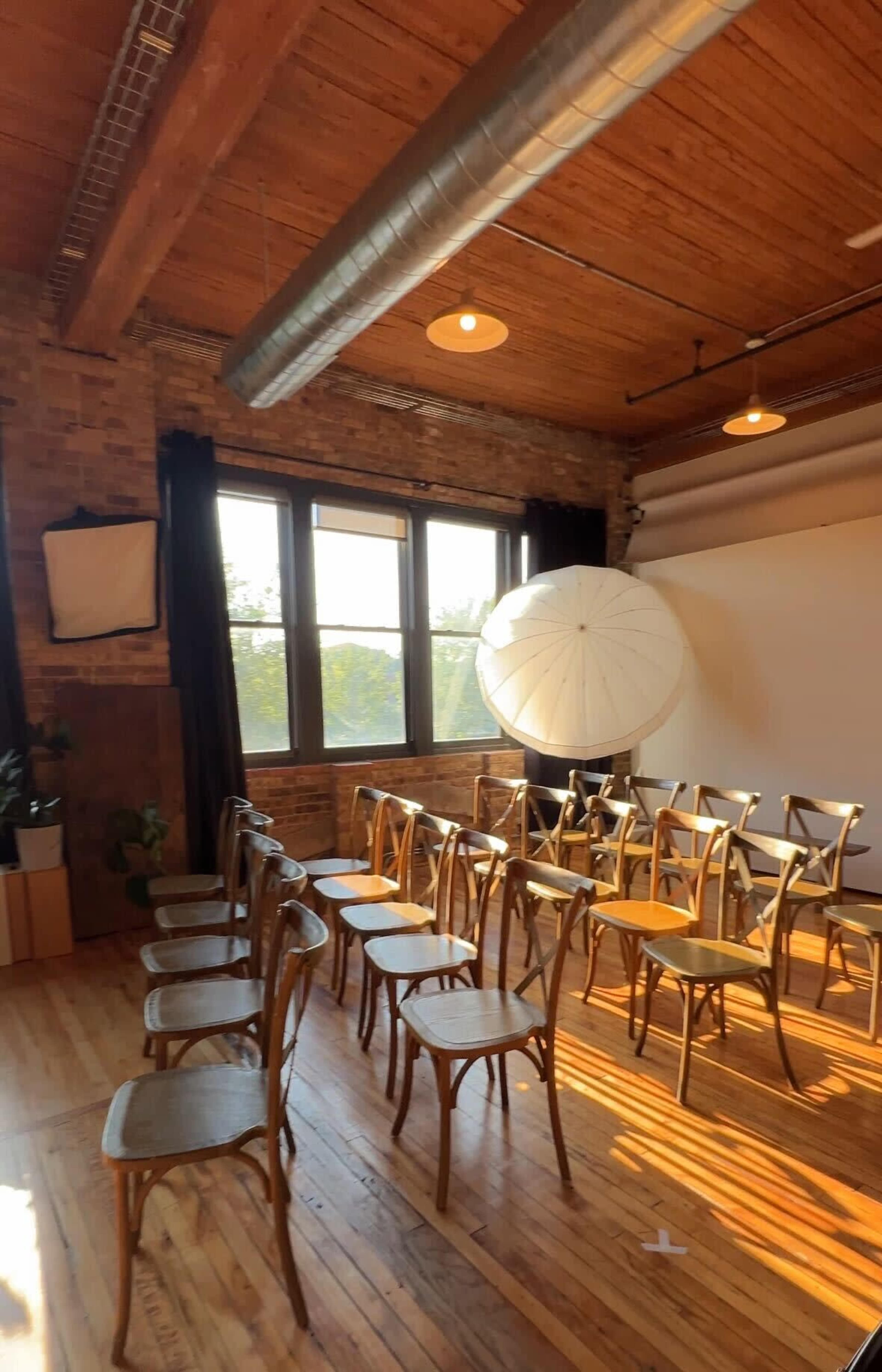 A row of wooden chairs is arranged in a sunlit room with exposed brick walls and large windows, featuring a large white umbrella in the corner.