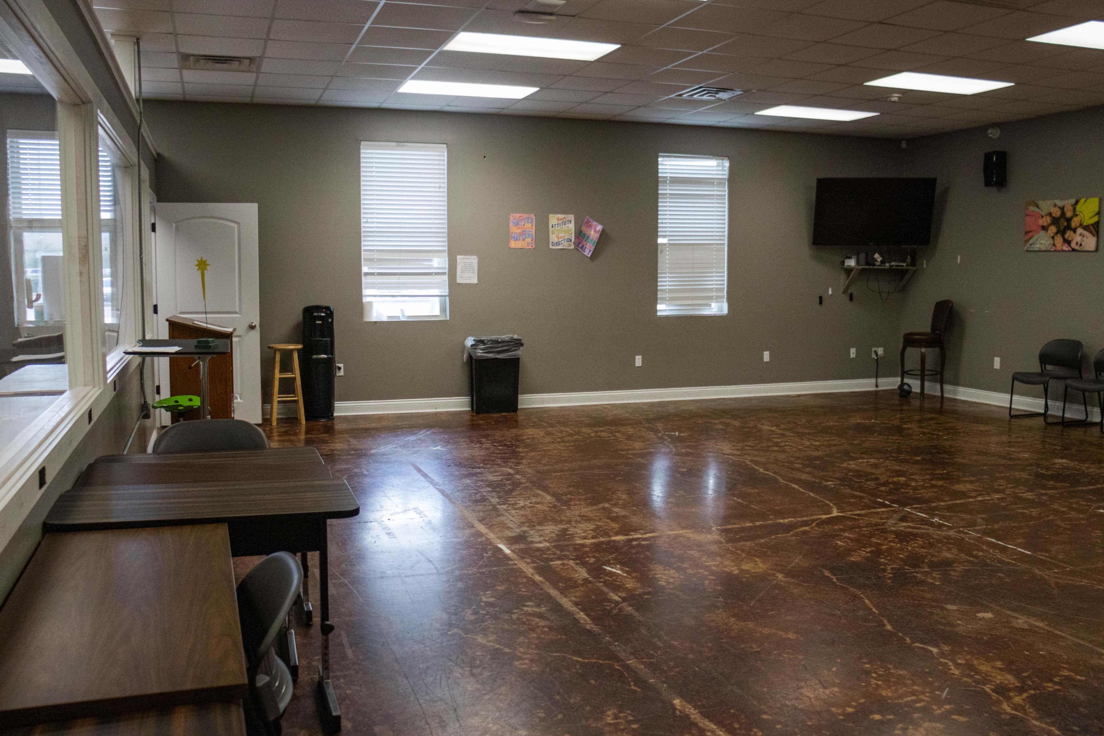 The image shows an empty room with a polished floor, several windows, a TV mounted on the wall, a trash can, and a few chairs.