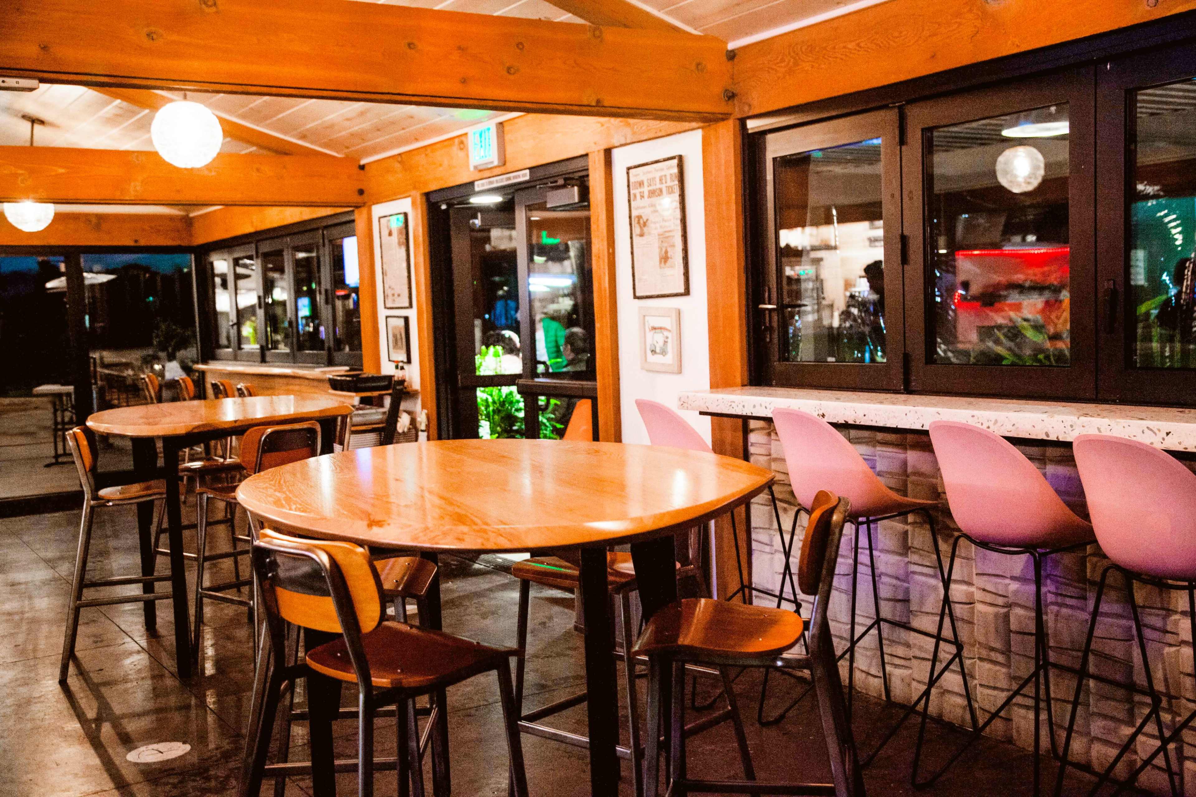 The image shows a dimly lit restaurant interior with wooden tables and pink bar stools set against large windows.