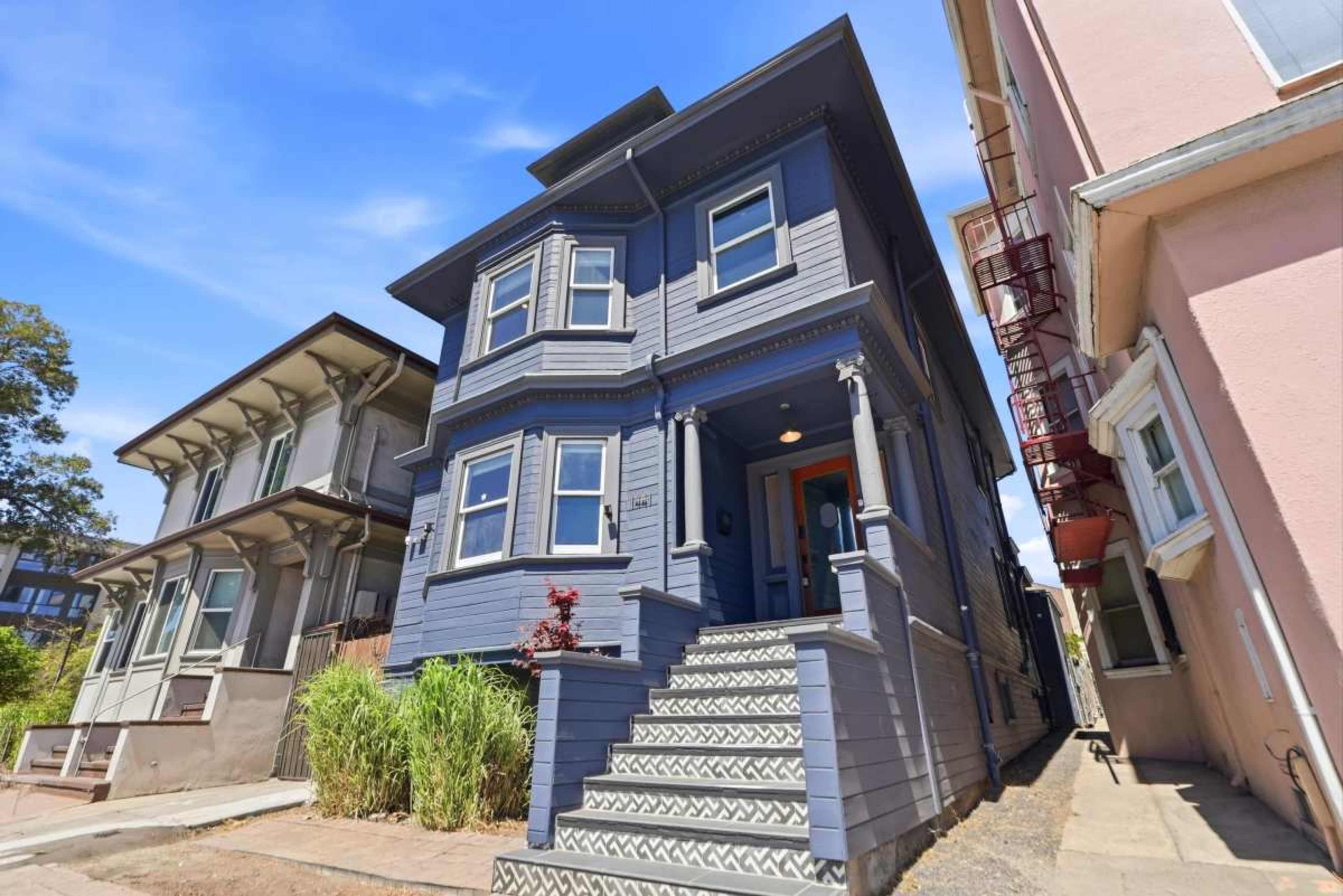A blue Victorian-style house stands at the corner of a street, flanked by two other buildings with differing architectural designs.
