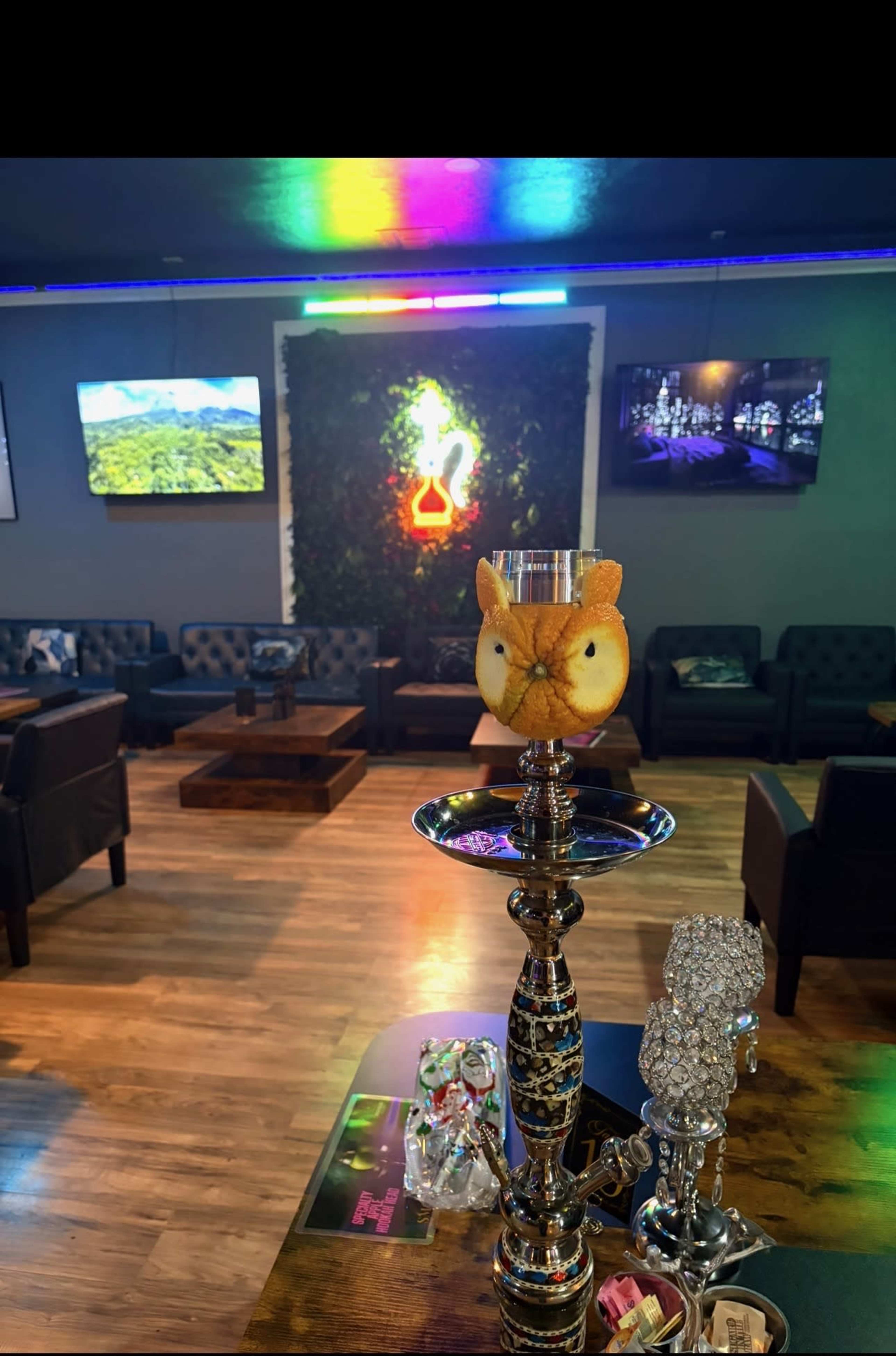 A hookah adorned with a decorative owl-shaped fruit sits on a table in a modern lounge with plush seating and colorful lighting.