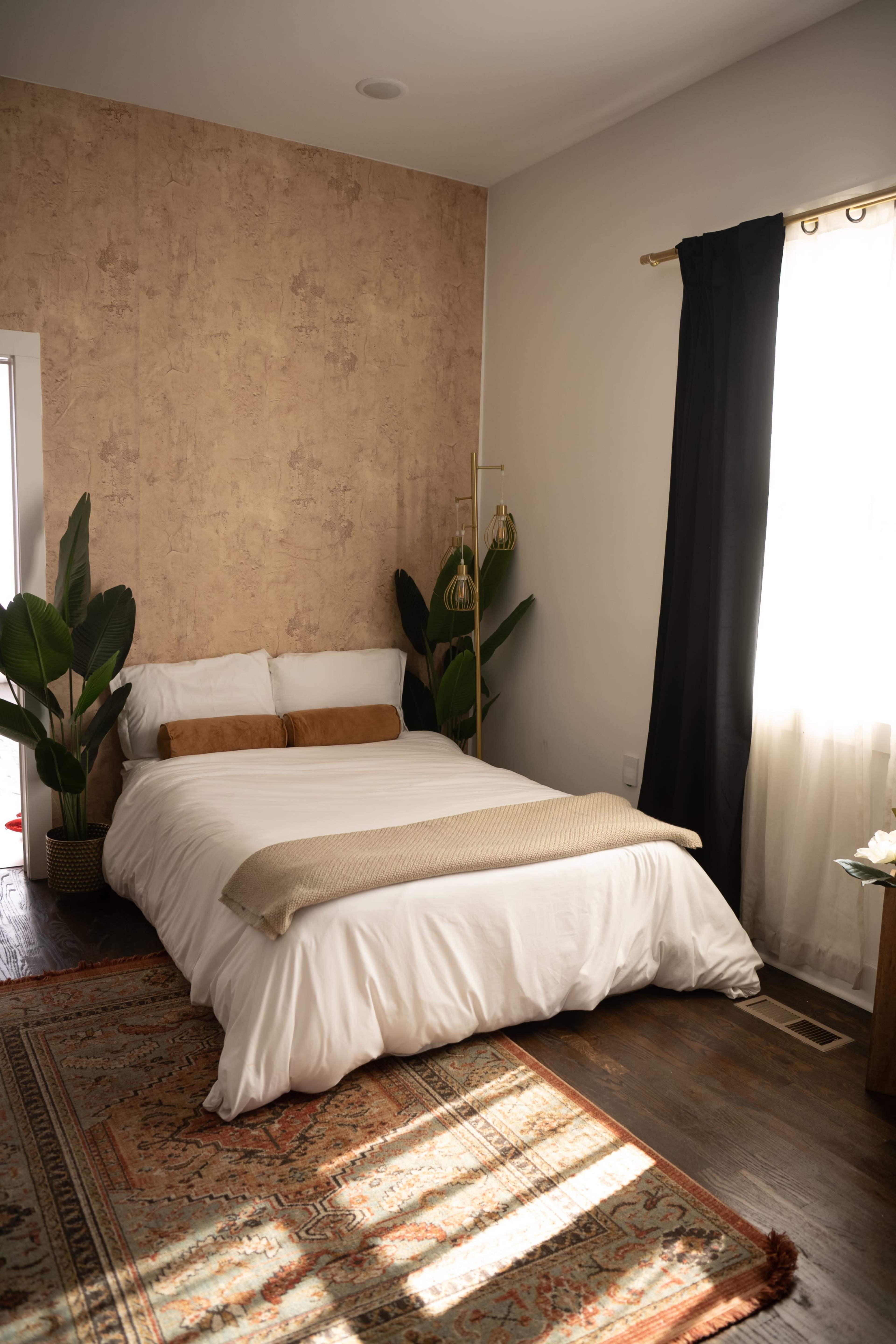 The image shows a cozy bedroom featuring a bed with white bedding, a patterned area rug, and plants beside the bed, illuminated by natural light coming through a window.