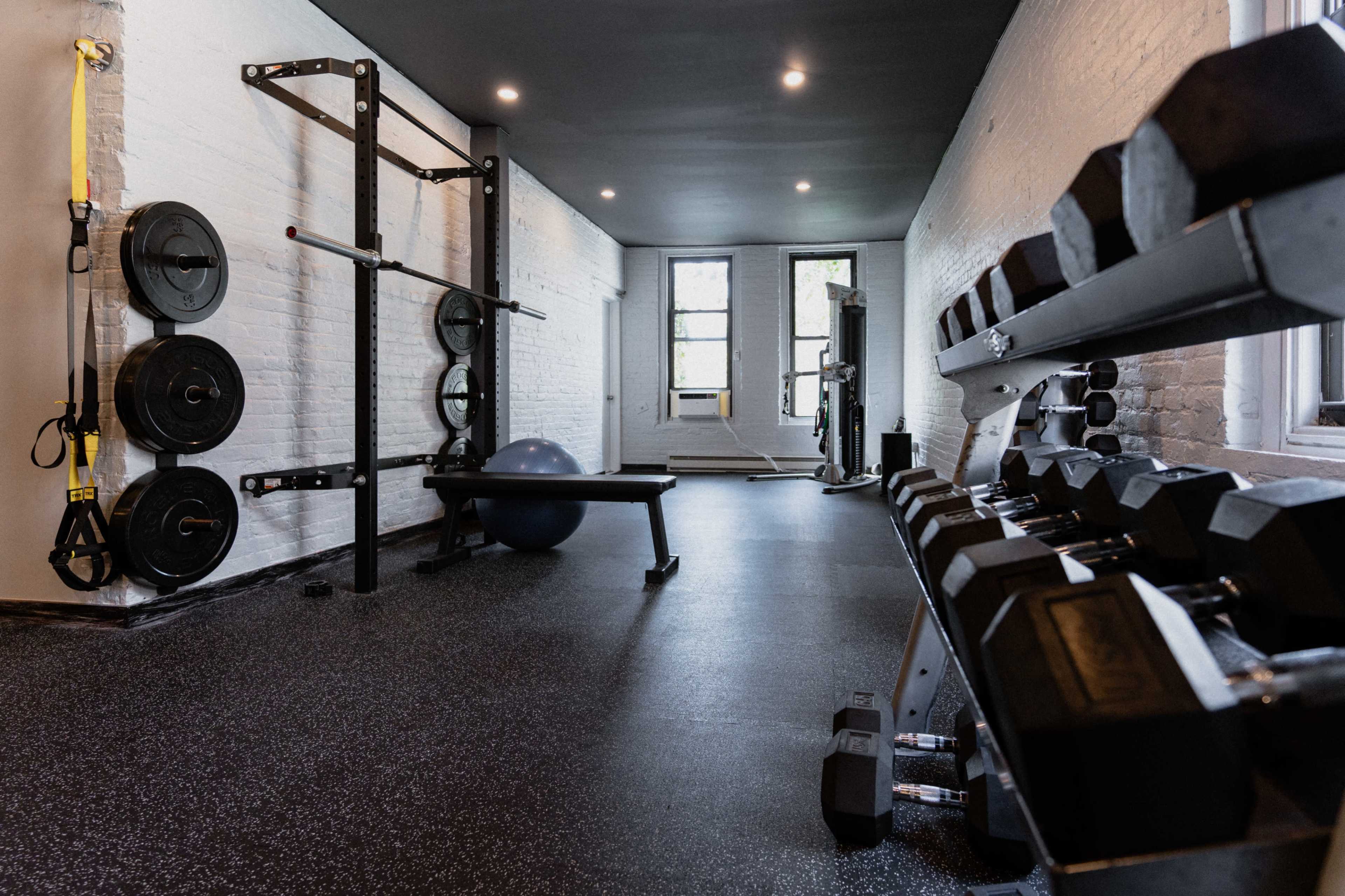 A well-equipped gym features a range of free weights, a bench, resistance bands, and a large exercise ball in a spacious room with white brick walls and windows.