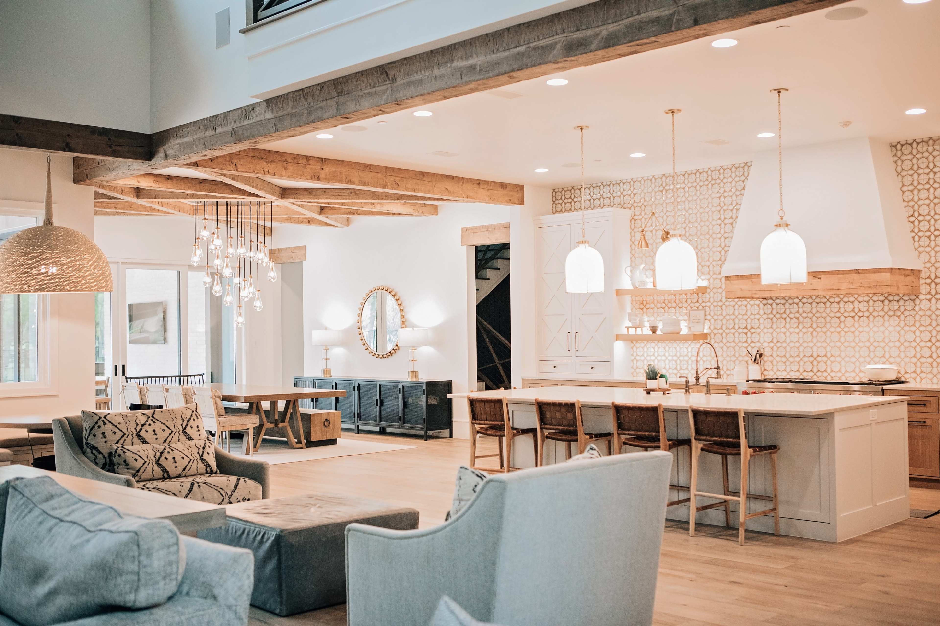 A modern open-concept living space with a kitchen island, dining area, and comfortable seating arrangements, featuring wooden beams and stylish light fixtures.