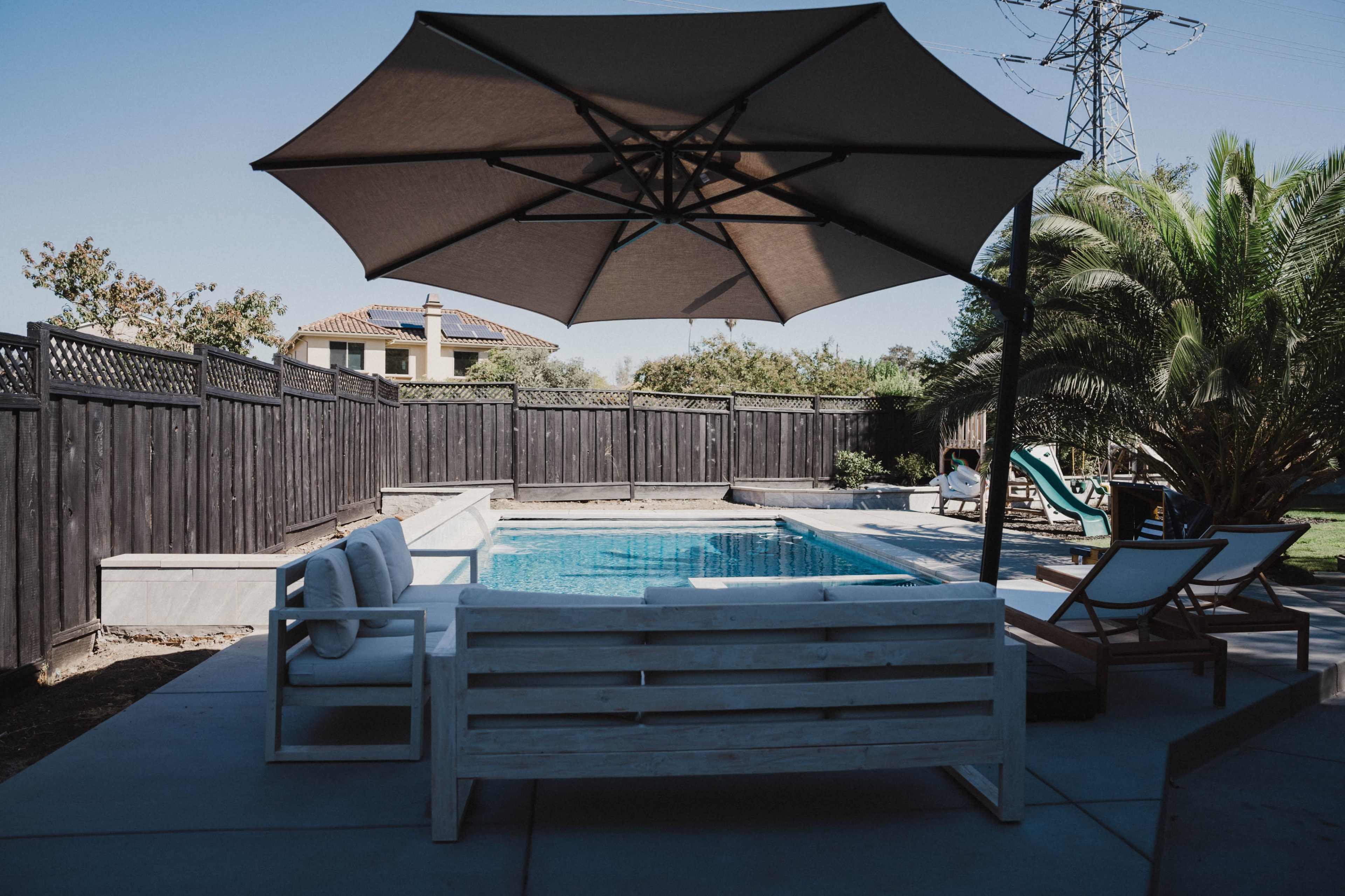 A shaded seating area is positioned beside a swimming pool, surrounded by a black wooden fence and landscaping.