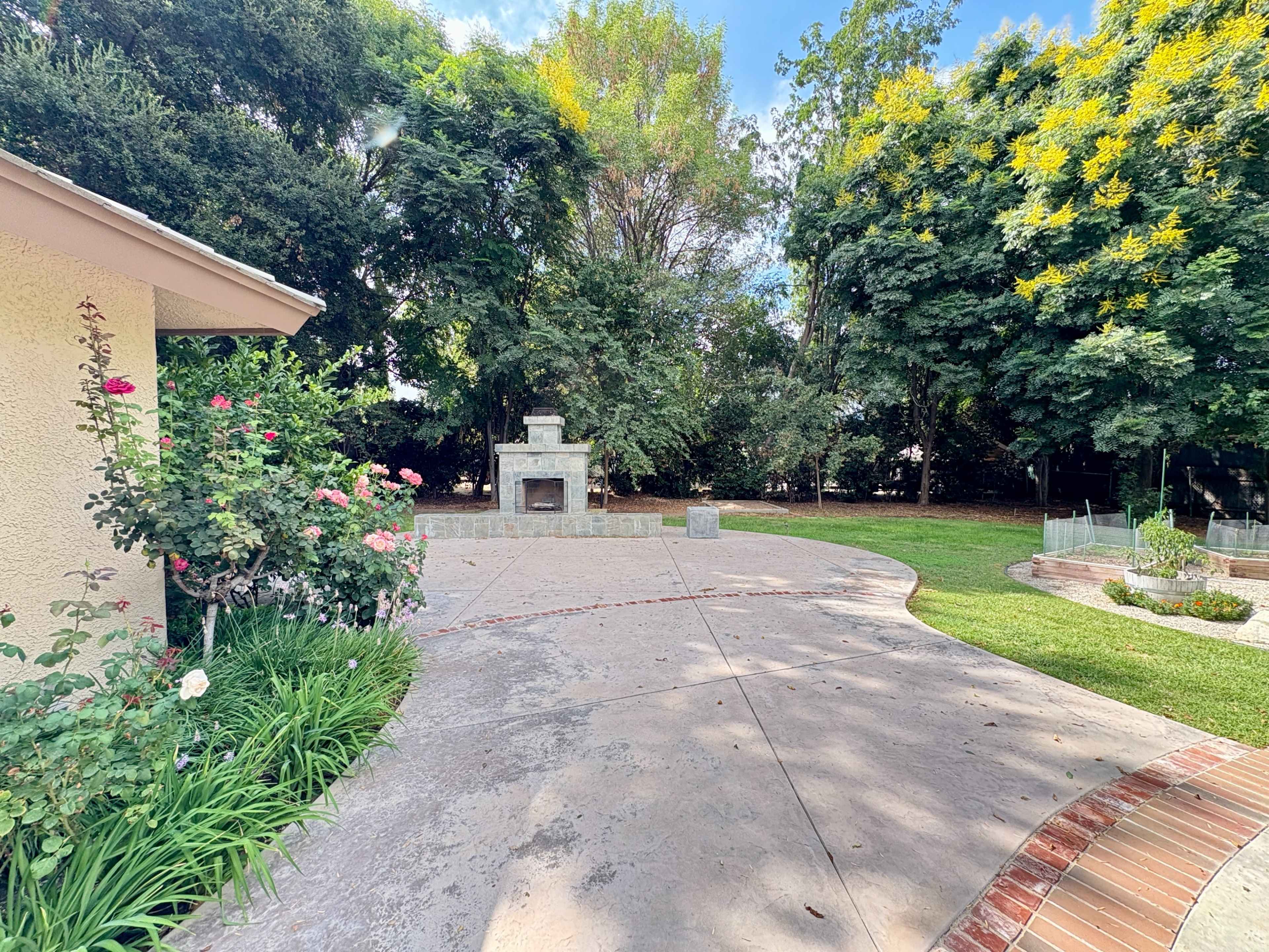 The image shows a landscaped backyard with a stone fireplace, surrounded by trees and flowering plants.