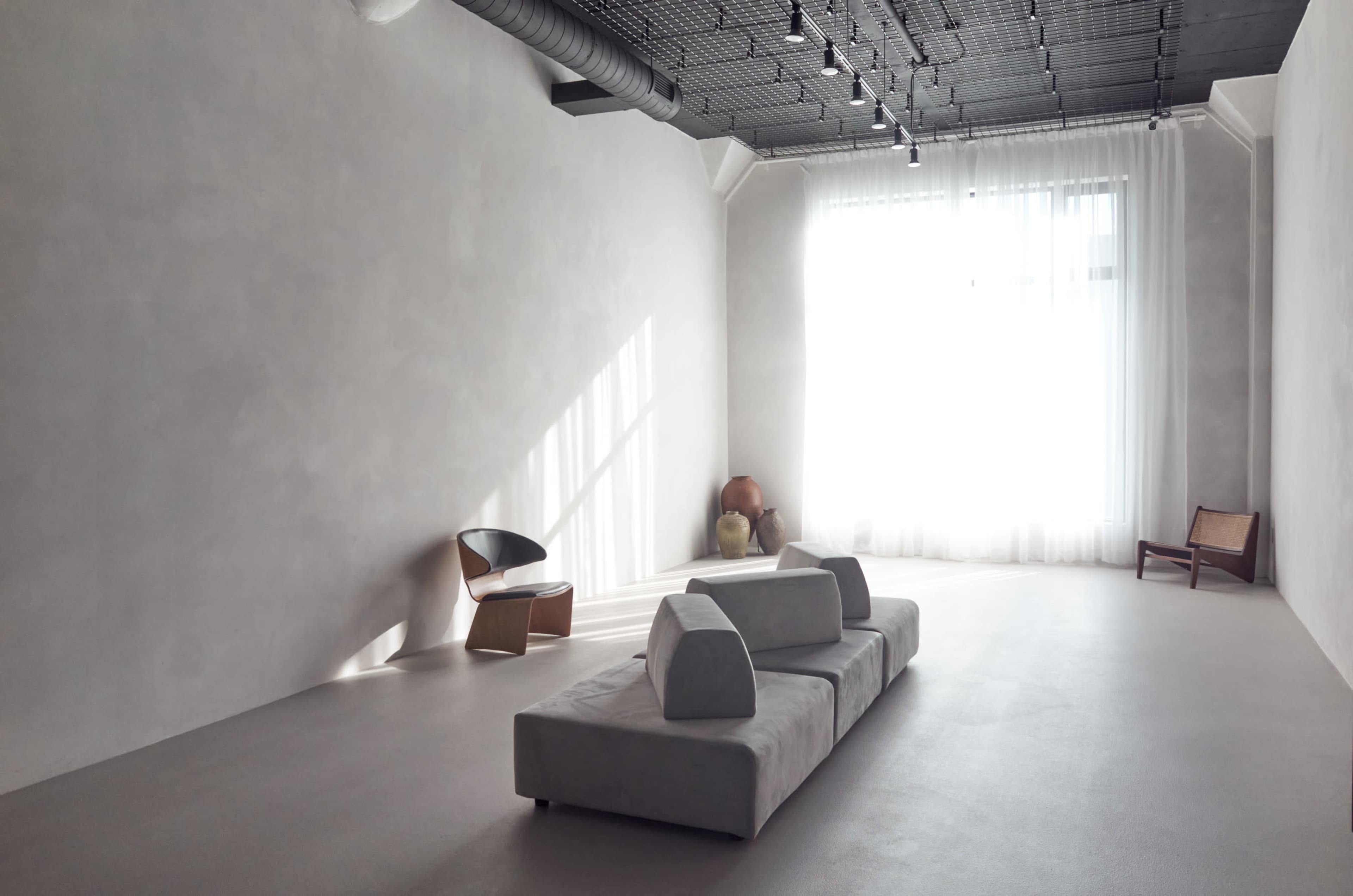 The image shows a minimalist interior space with a light-colored sofa, wooden chairs, and natural light streaming through large windows.