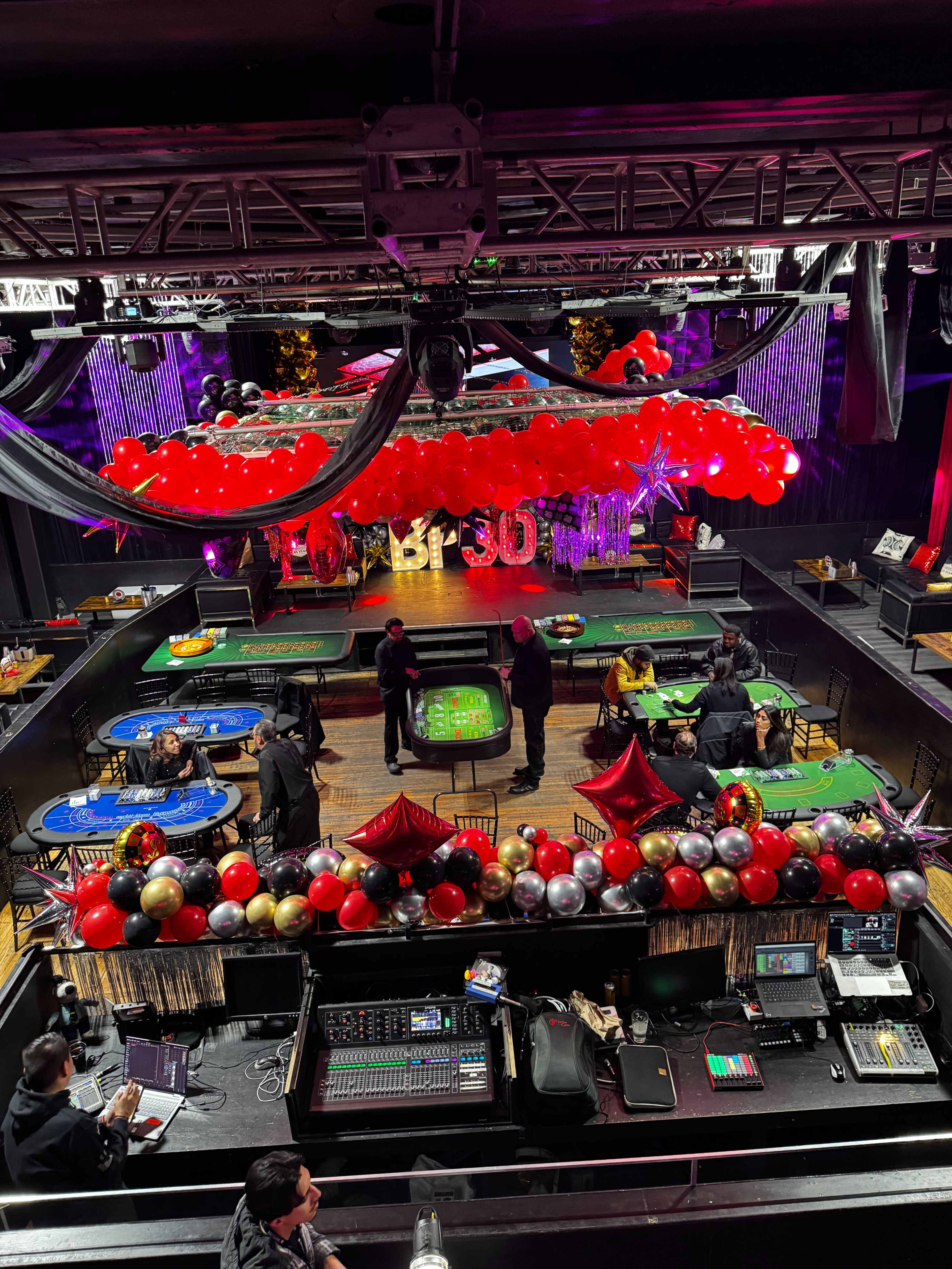 The image shows a vibrant casino-themed event space with gaming tables, decorative balloons, and a DJ setup.