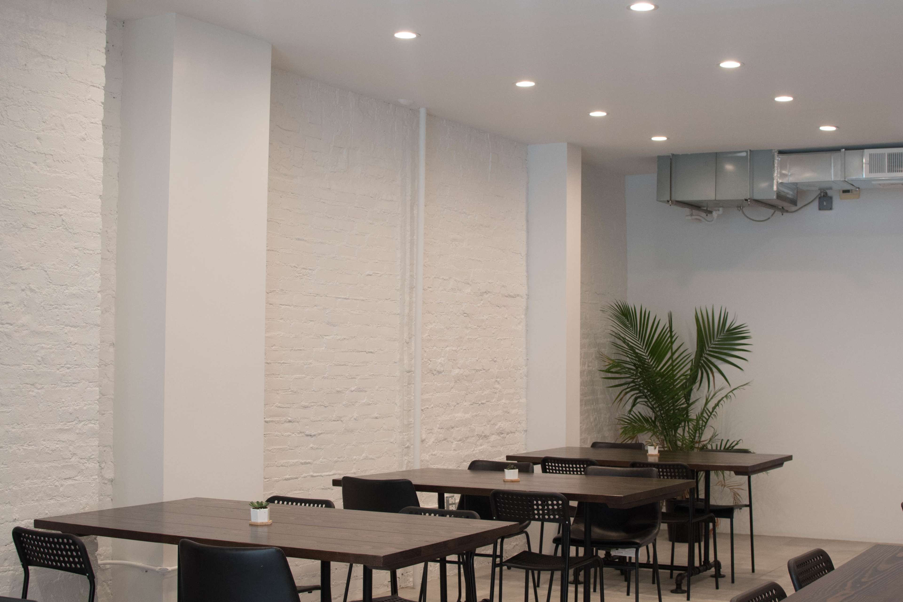 A minimalist indoor dining area with several wooden tables and black chairs, along with a potted plant in the corner.