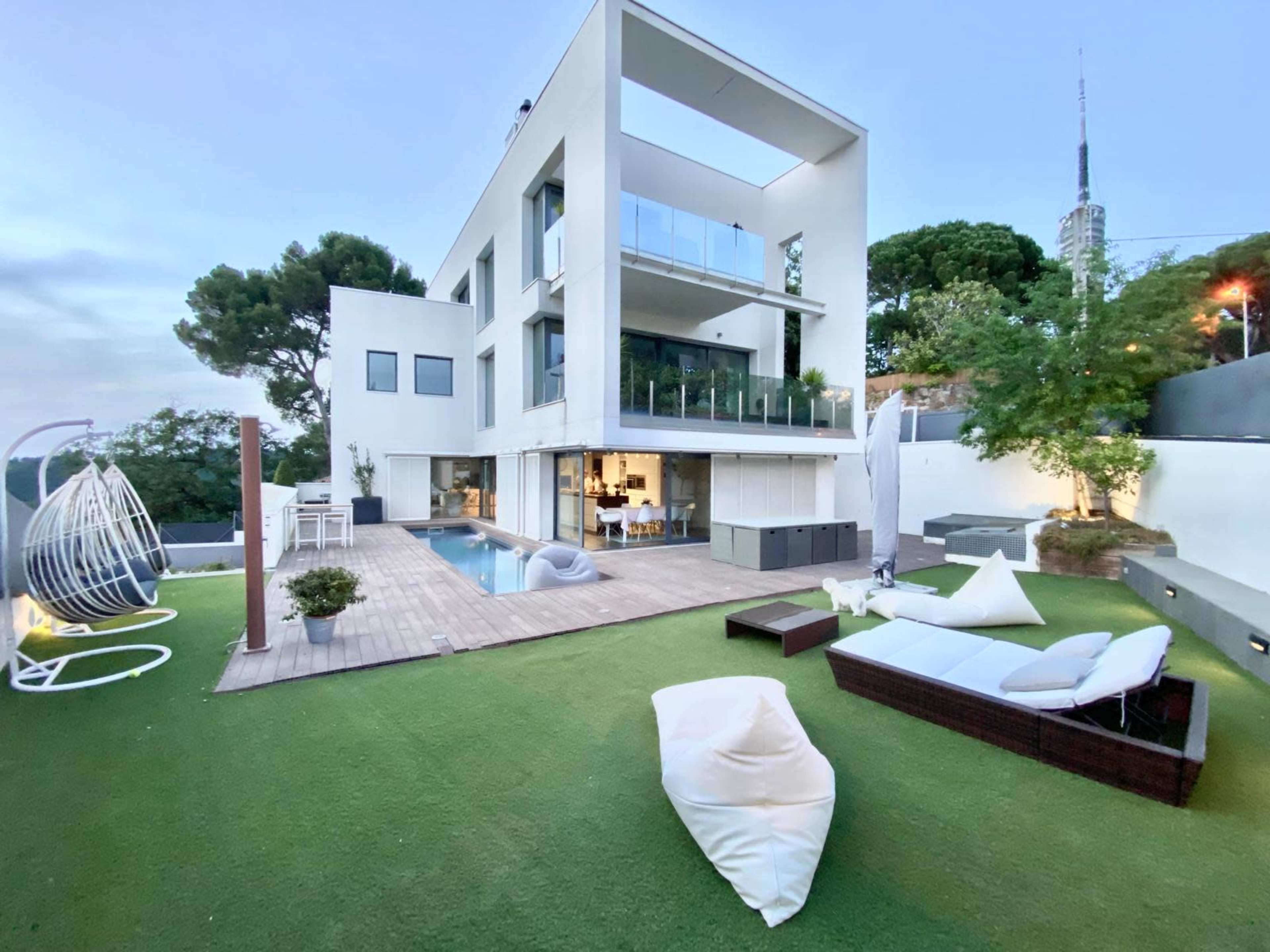 A modern two-story house with large windows overlooks a manicured lawn, featuring a swimming pool, outdoor seating areas, and a contemporary design.