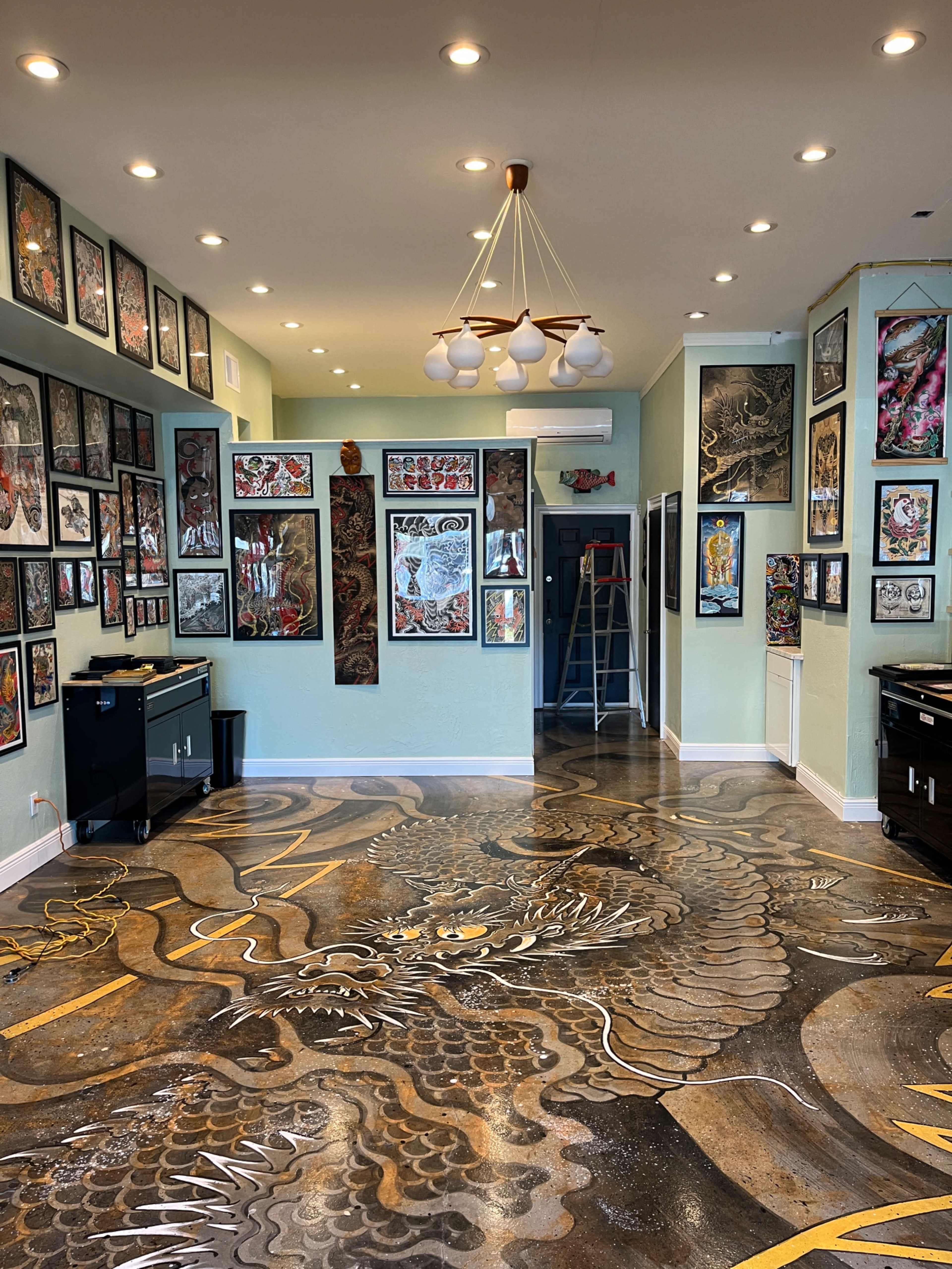 The image depicts a tattoo studio featuring a dragon-themed floor design, adorned with numerous framed artworks on the walls and various tools and equipment placed around the space.