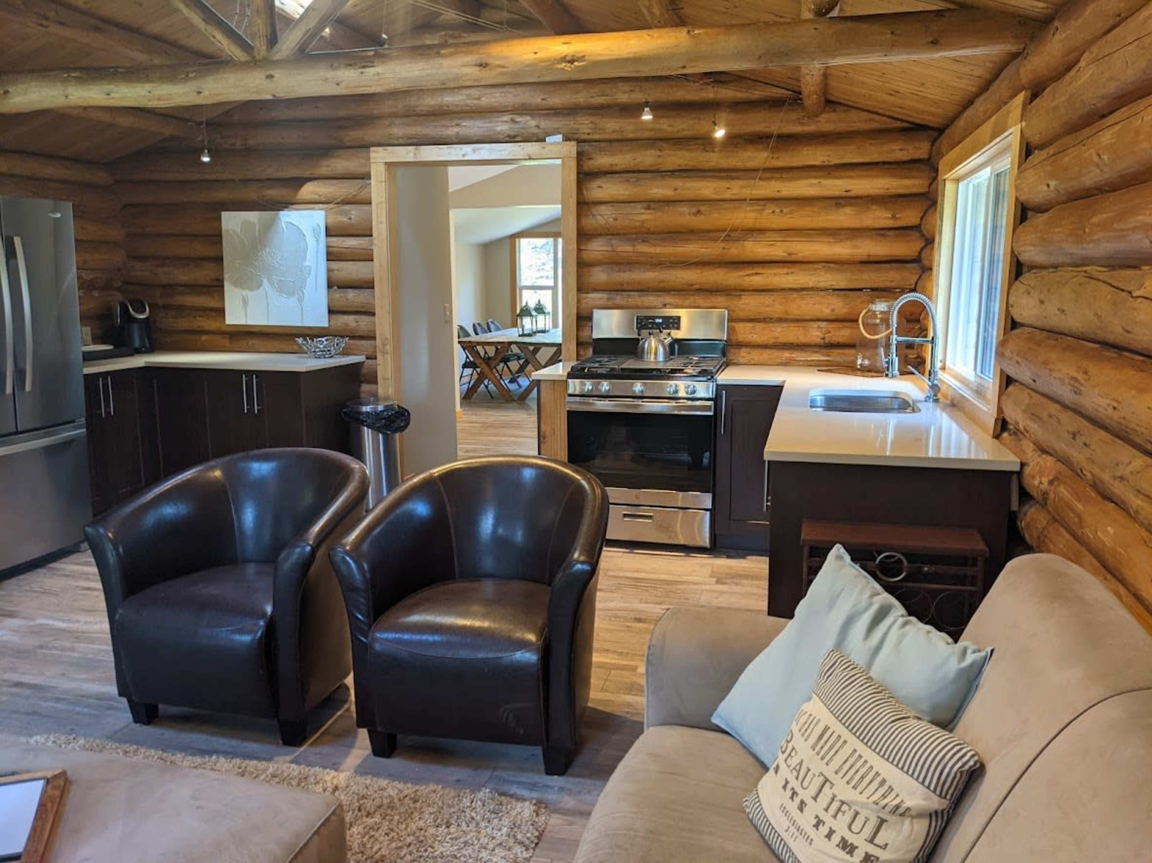 The interior of a log cabin features a kitchen with modern appliances, a dining area, and two leather chairs facing a sofa.