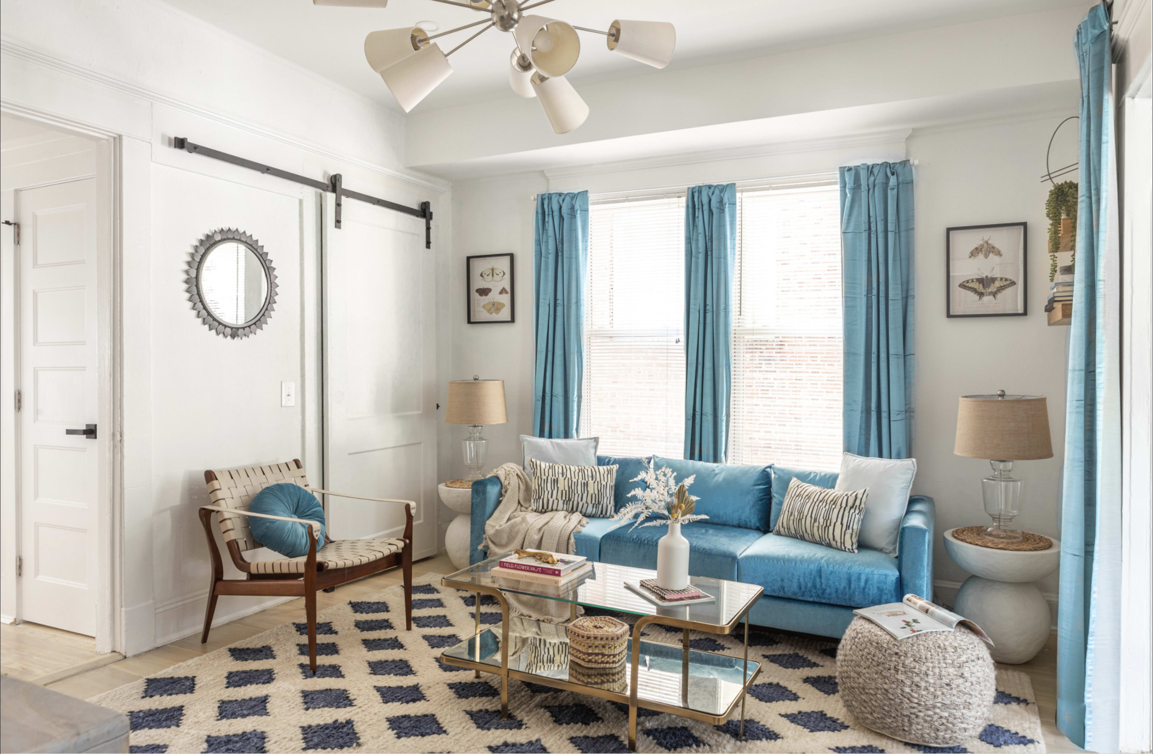 The image shows a bright, modern living room with a blue sofa, a coffee table, and large windows with light blue curtains.