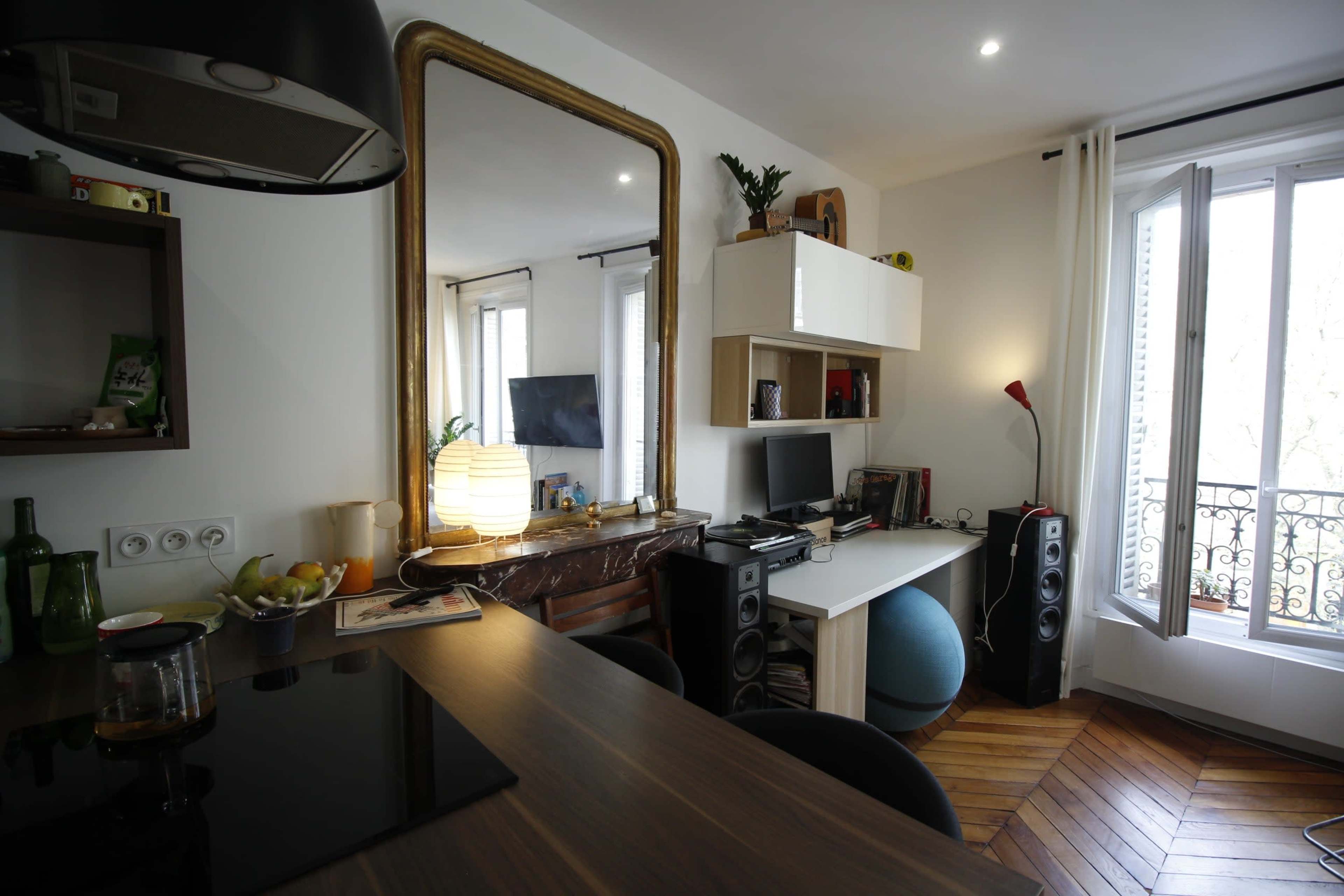 A modern, compact living space featuring a kitchen area, a desk with a computer, and a large mirror, adorned with a few decorative plants.