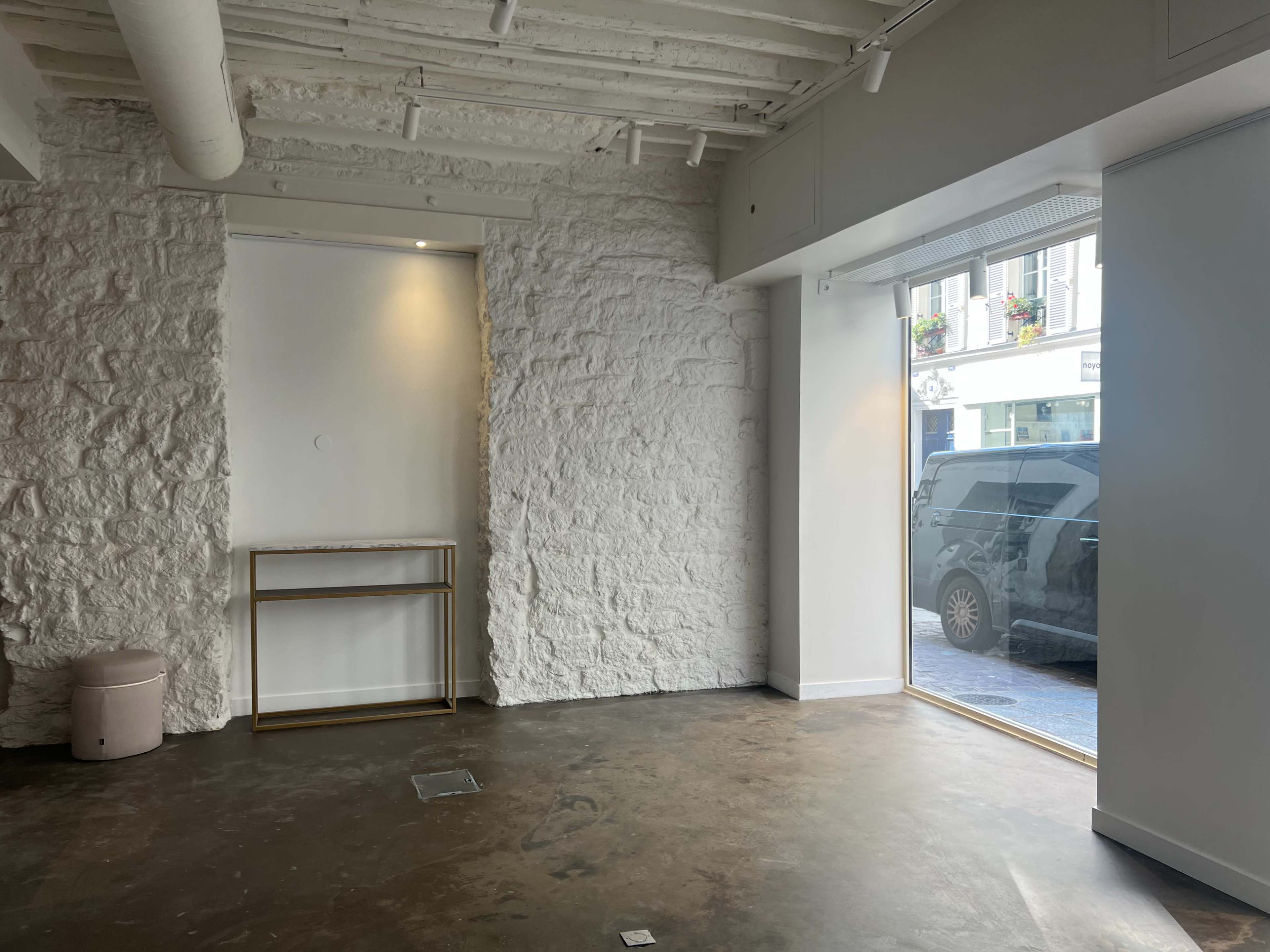 The image shows a modern, empty room with a textured white wall, a small gold-framed table, and an open glass door leading to a street.