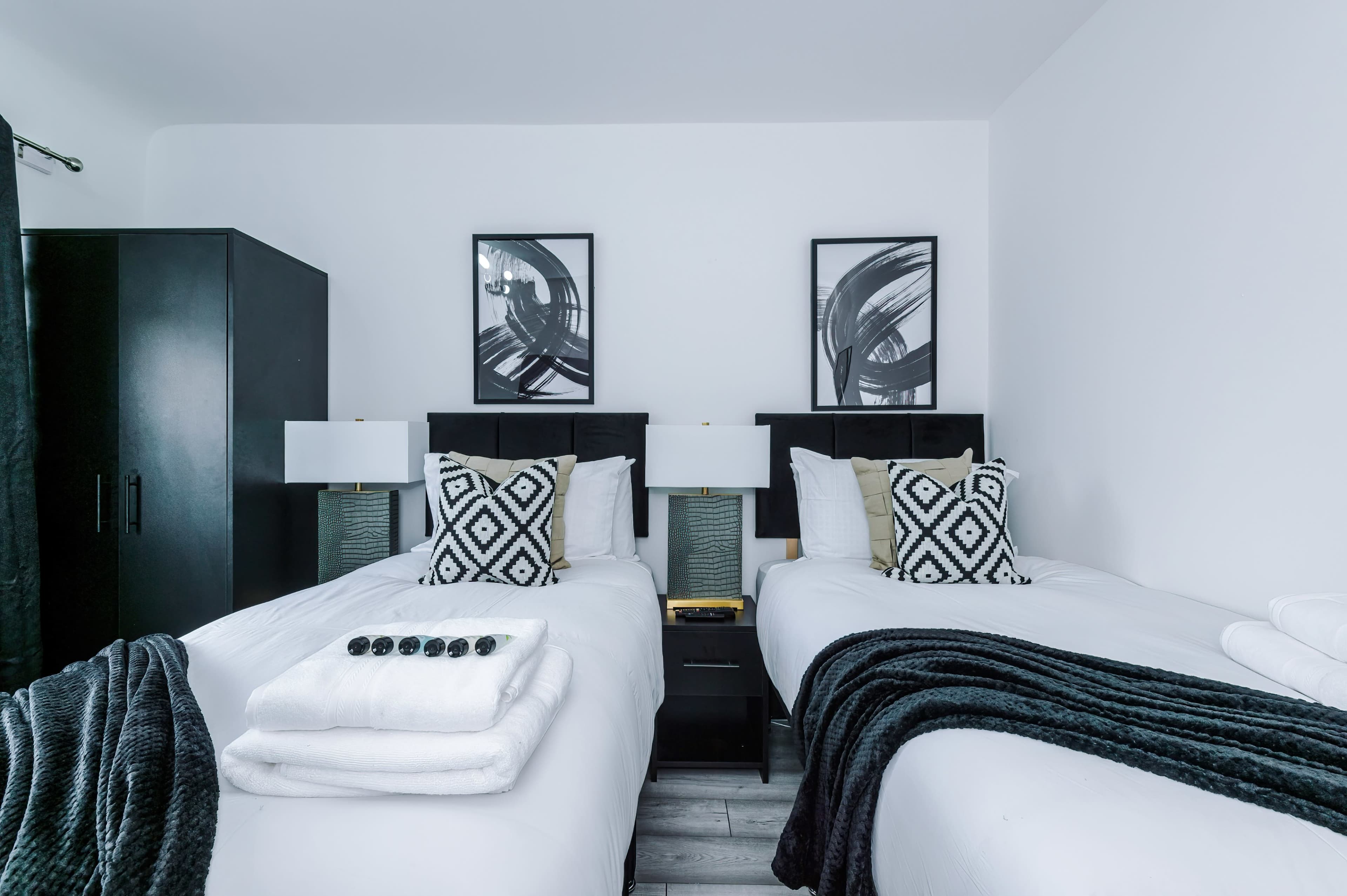 A modern hotel room with two twin beds, black accent furniture, and artwork on the walls.