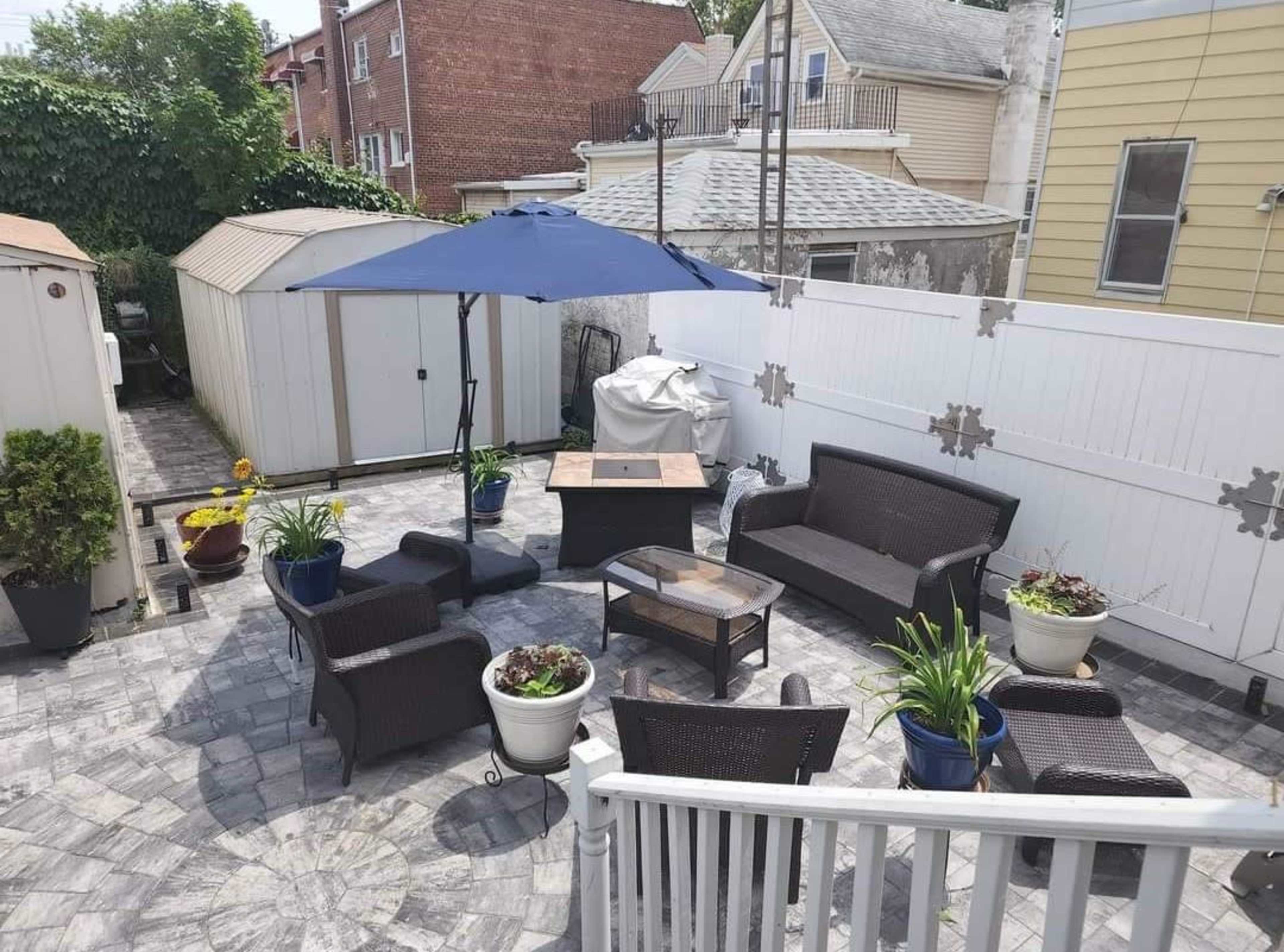 A backyard patio area with a seating arrangement around a fire pit, surrounded by potted plants and a large umbrella.