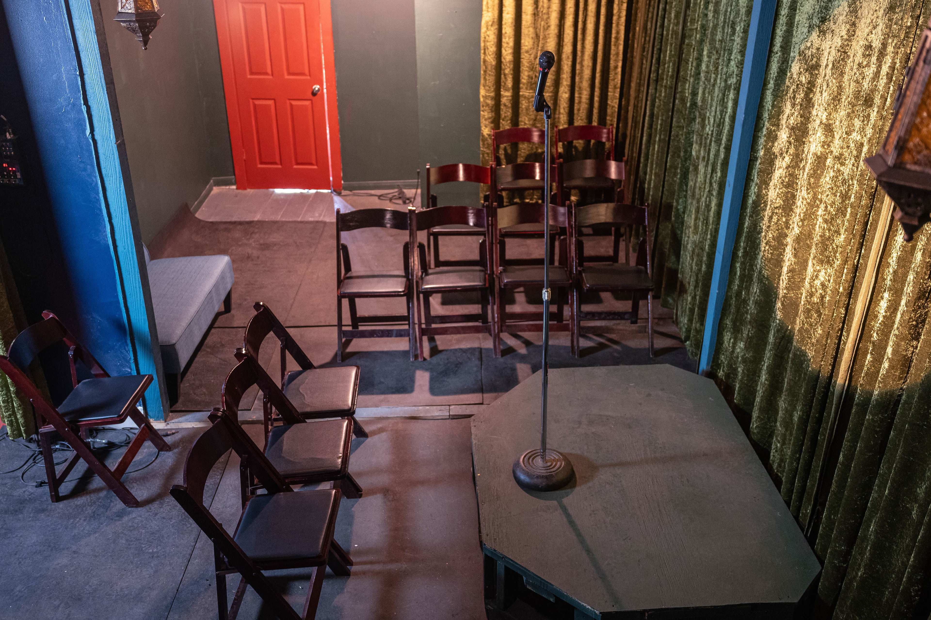 A small stage with a microphone on a podium, surrounded by empty folding chairs and a red door in the background.