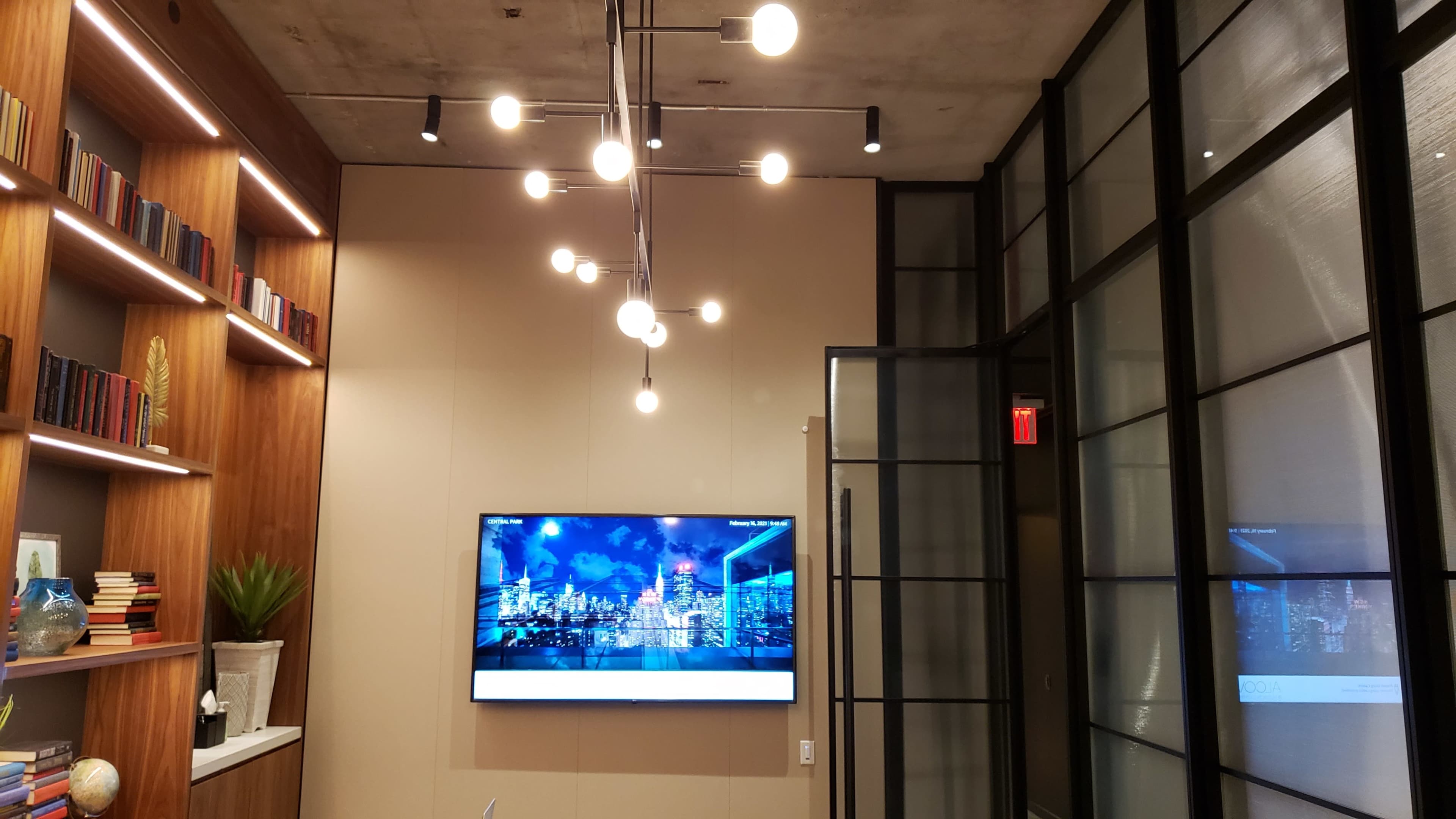 The image shows a modern interior with a bookshelf on the left, a large television displaying a cityscape, and a unique light fixture hanging from the ceiling.