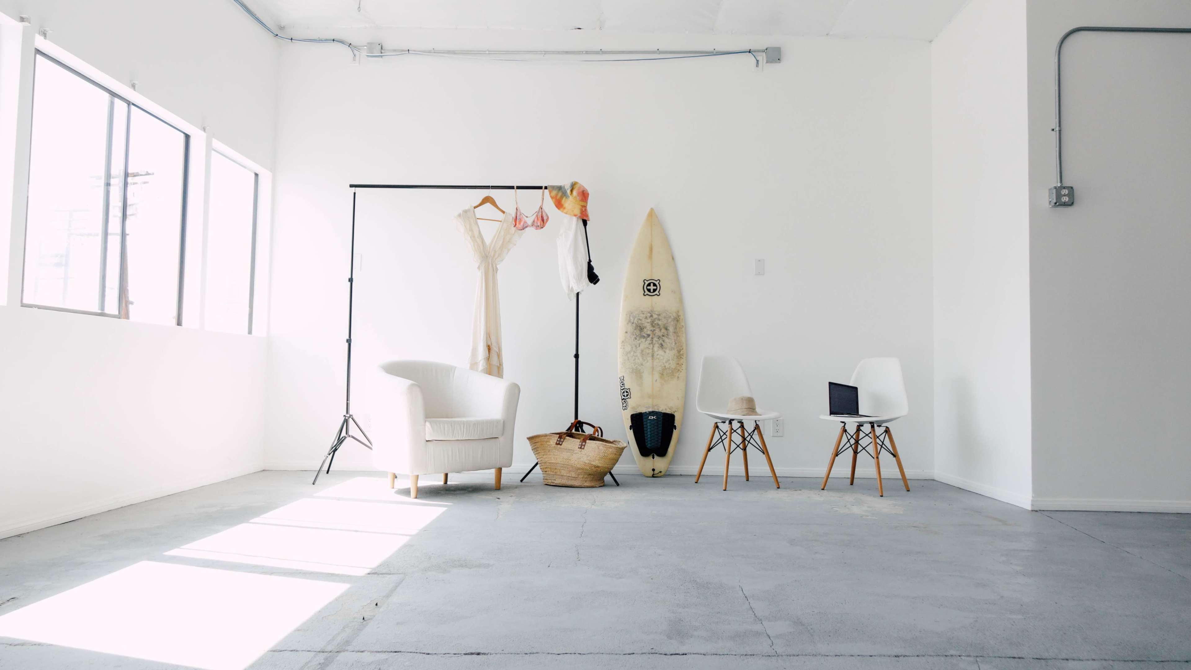 A minimalist interior space featuring a white chair, a clothing rack with a dress and accessories, a surfboard, a basket, and two chairs, illuminated by natural light.