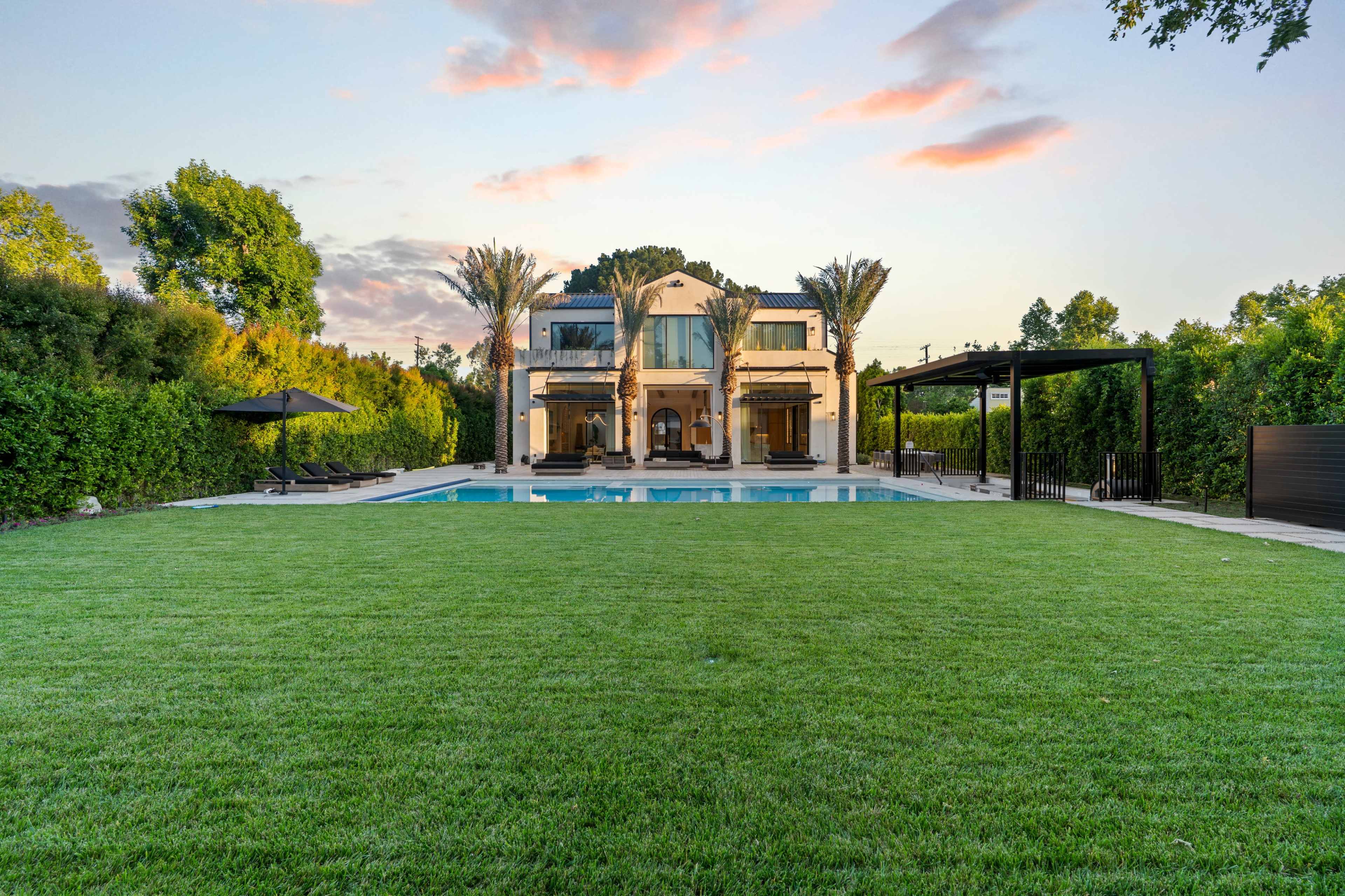 The image features a large, two-story villa with a swimming pool surrounded by a well-manicured lawn and tall palm trees.