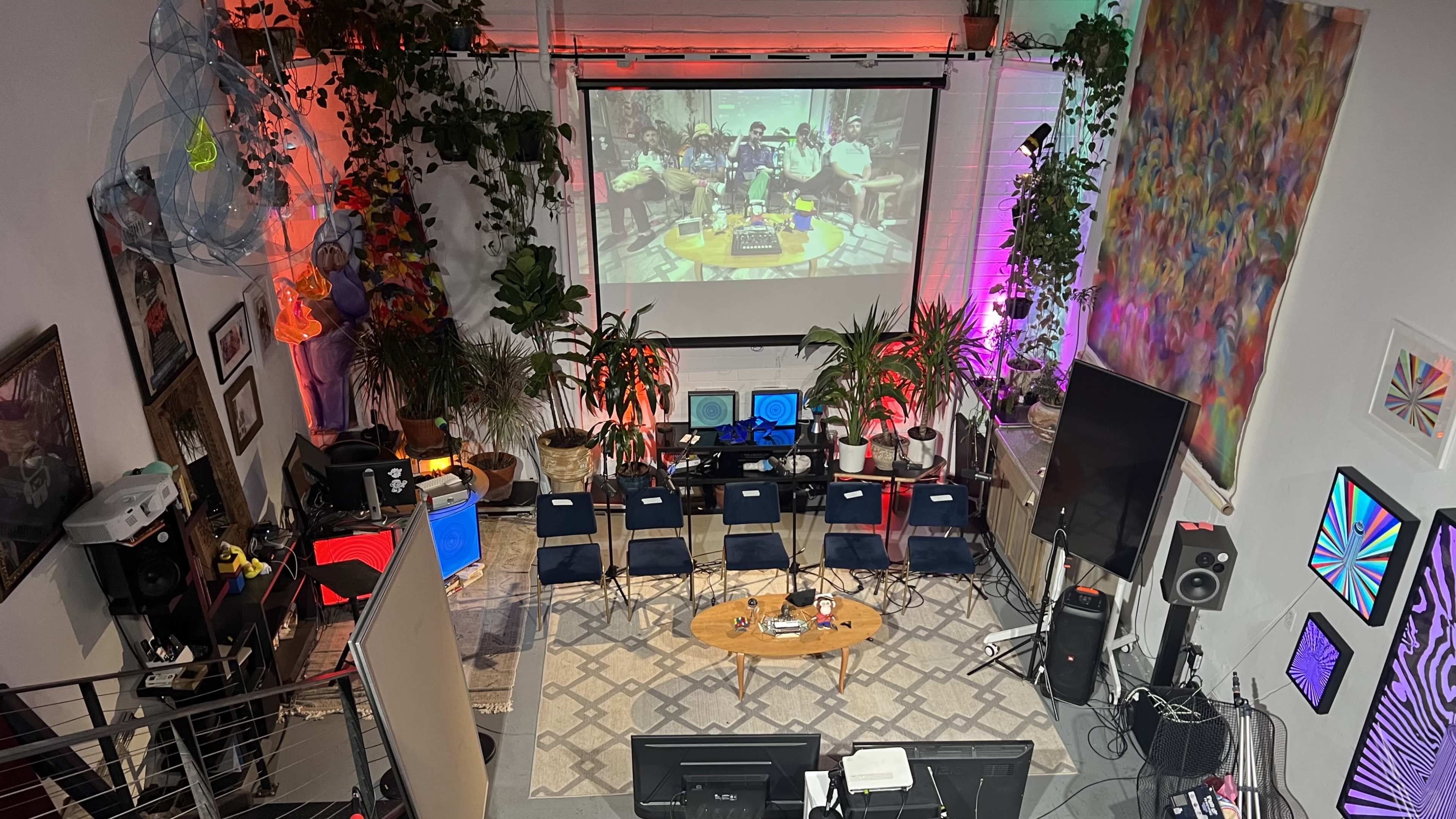 A vibrant indoor space set up with a large screen displaying a performance, surrounded by potted plants, chairs, and various audio equipment.