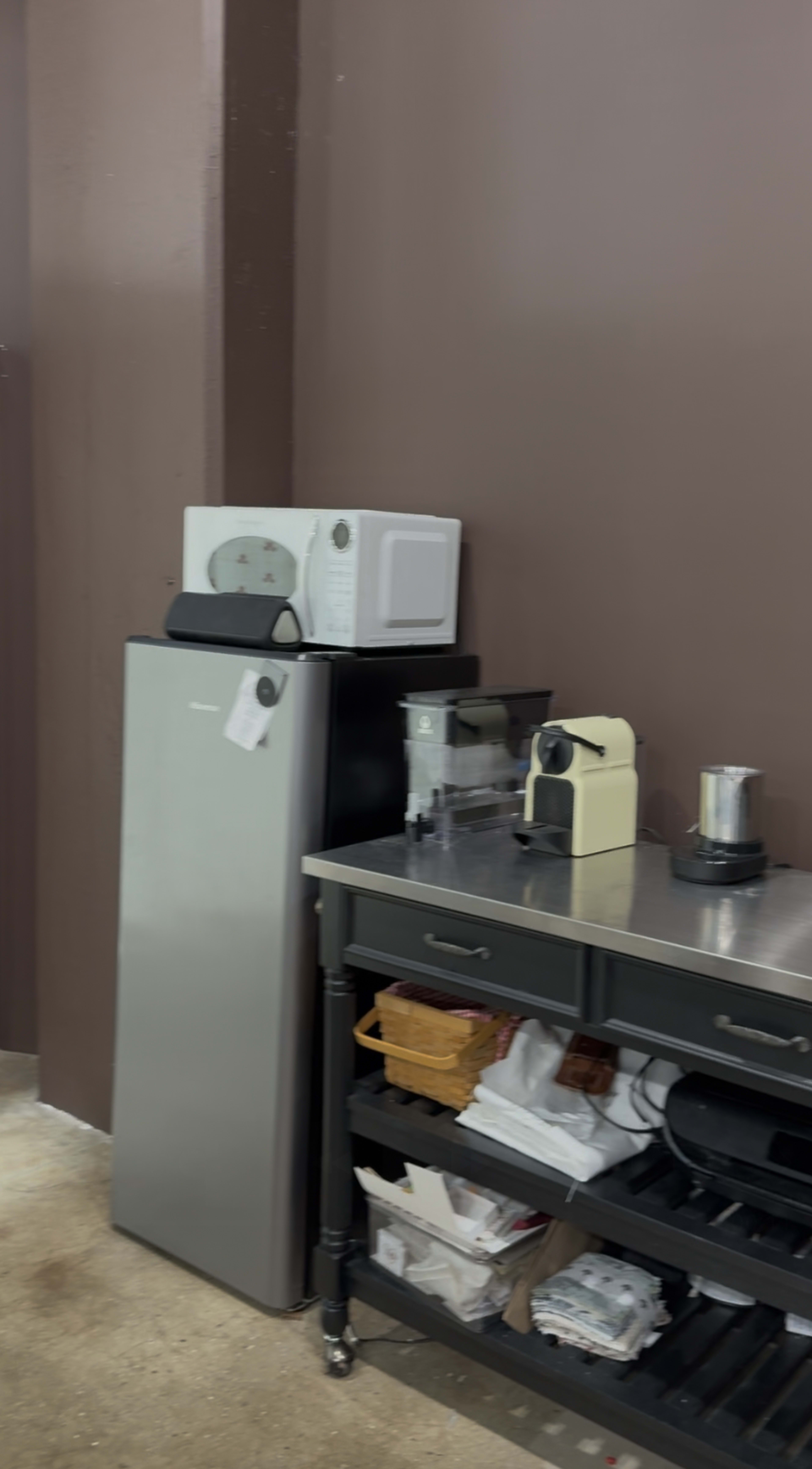 The image shows a kitchen corner featuring a silver refrigerator, a microwave, a coffee machine, and a stainless steel countertop with various kitchen items.