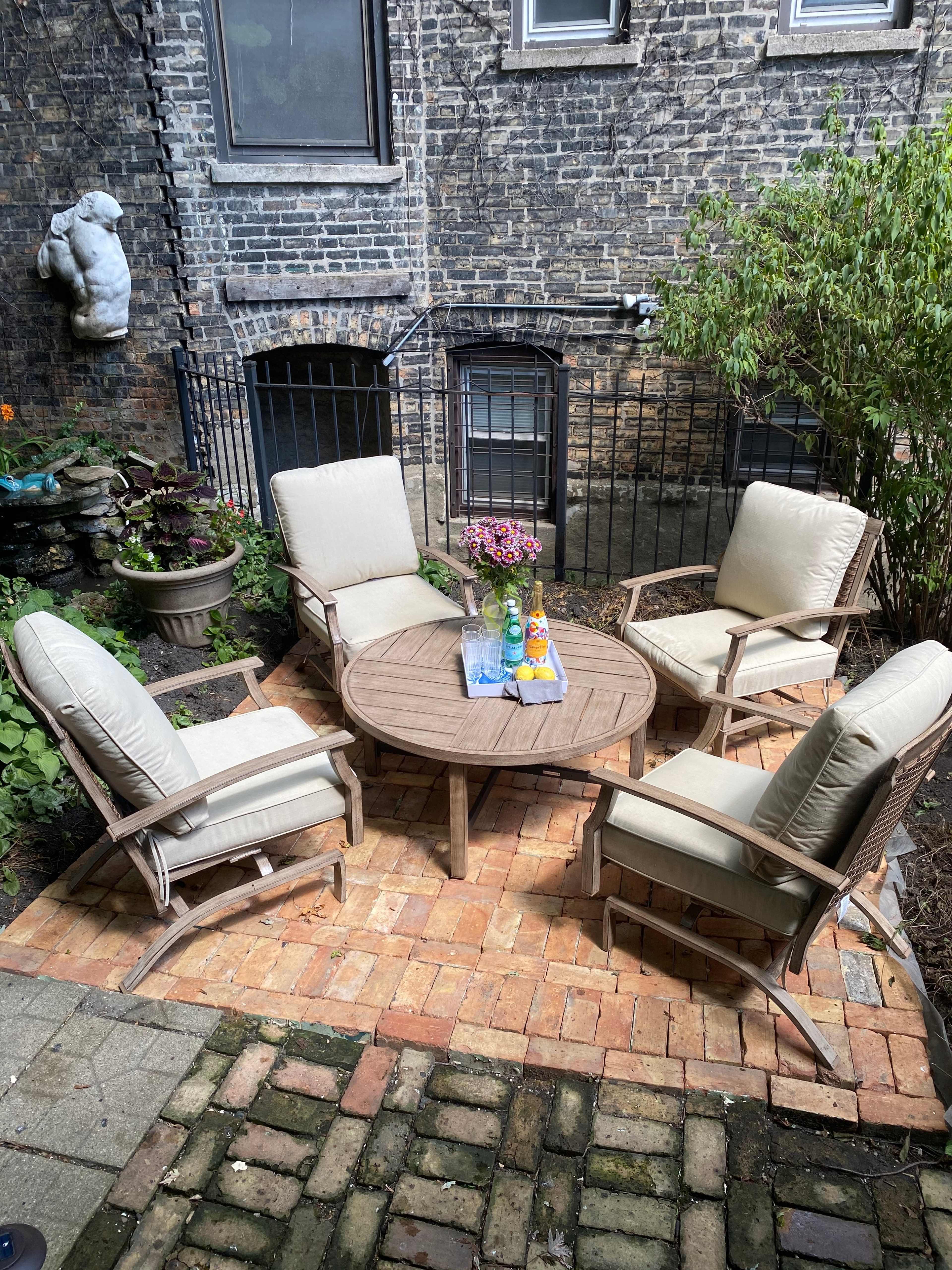 A circular wooden table surrounded by four cushioned chairs is set in a small brick-paved outdoor space with greenery and decorative elements.