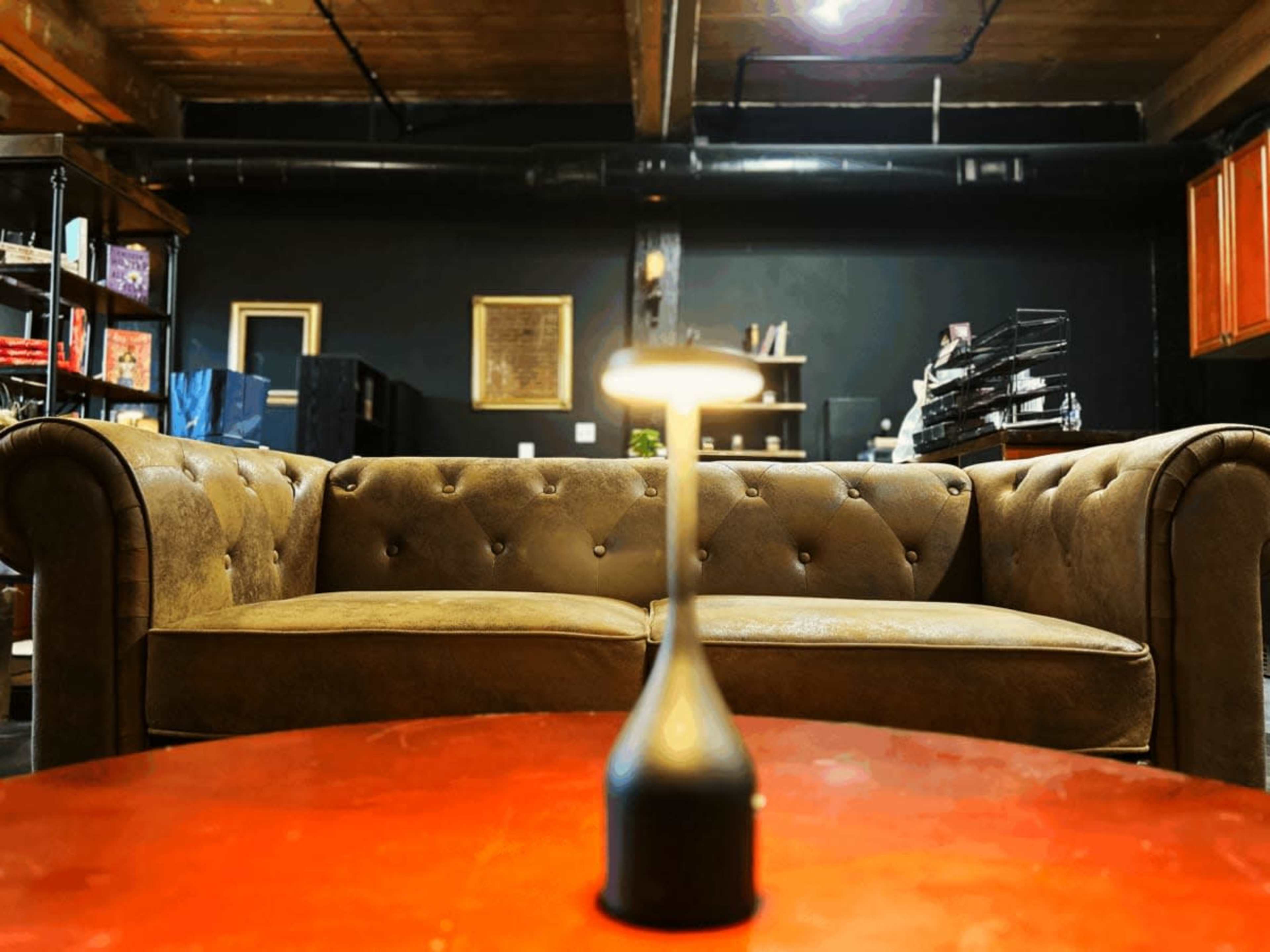 A brown leather couch is positioned in front of a low table with a lamp, set in a dimly lit room with dark walls and shelves in the background.