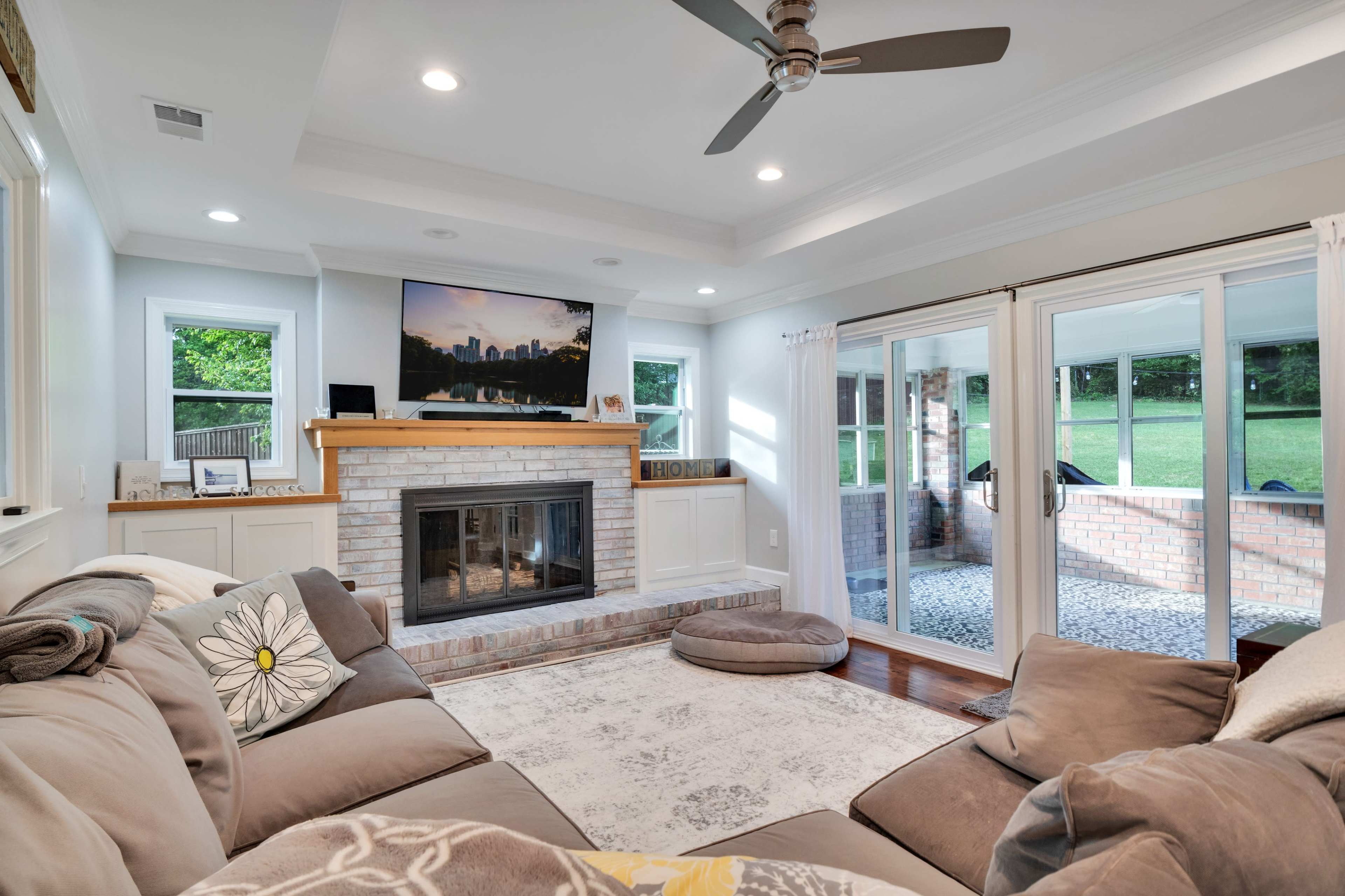 The living room features a cozy sectional sofa, a brick fireplace with a flat-screen TV above it, and large windows leading to an outdoor space.