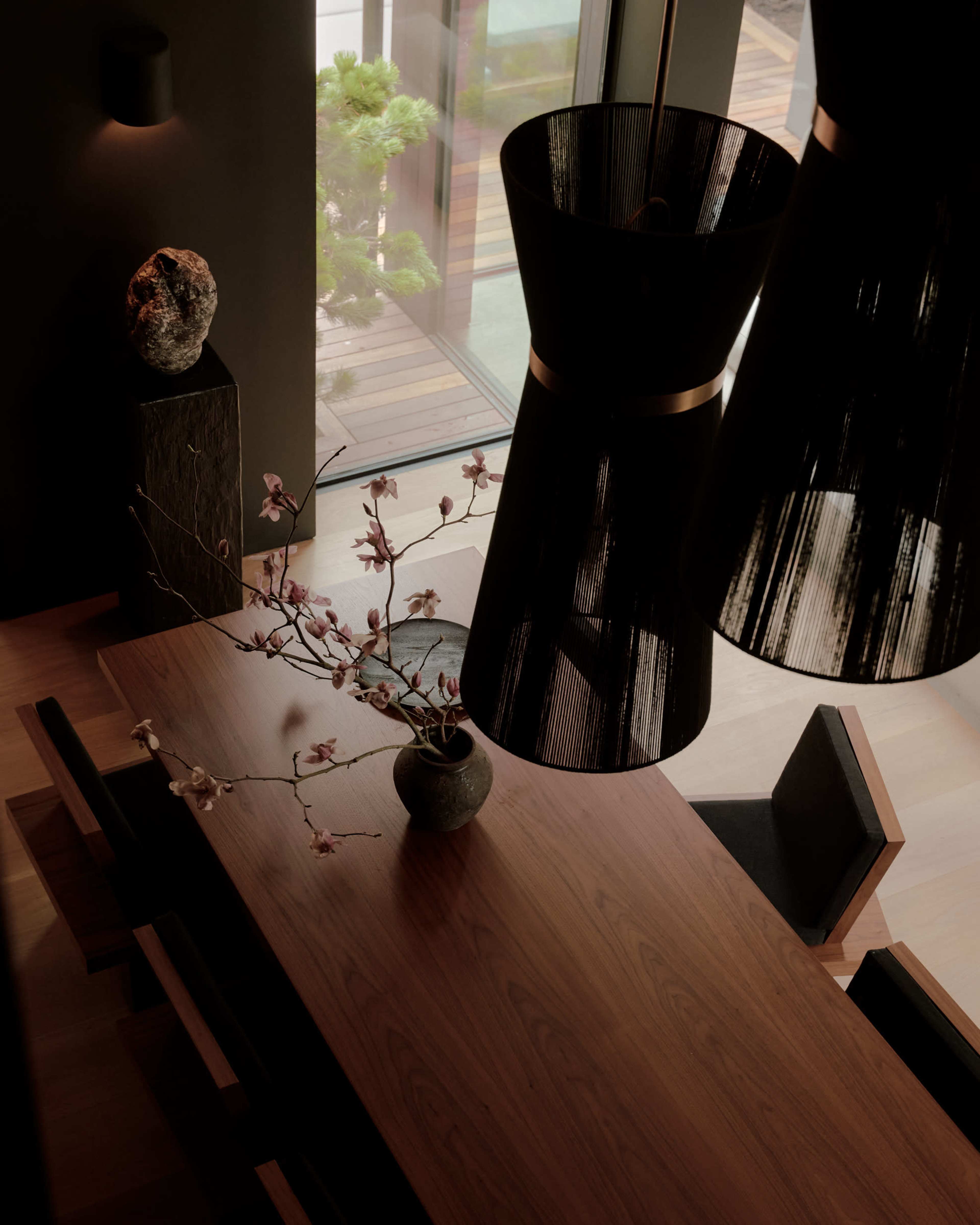 The image shows a modern dining room with a wooden table, a vase with a branch of flowers, and two large, dark pendant lights hanging above.