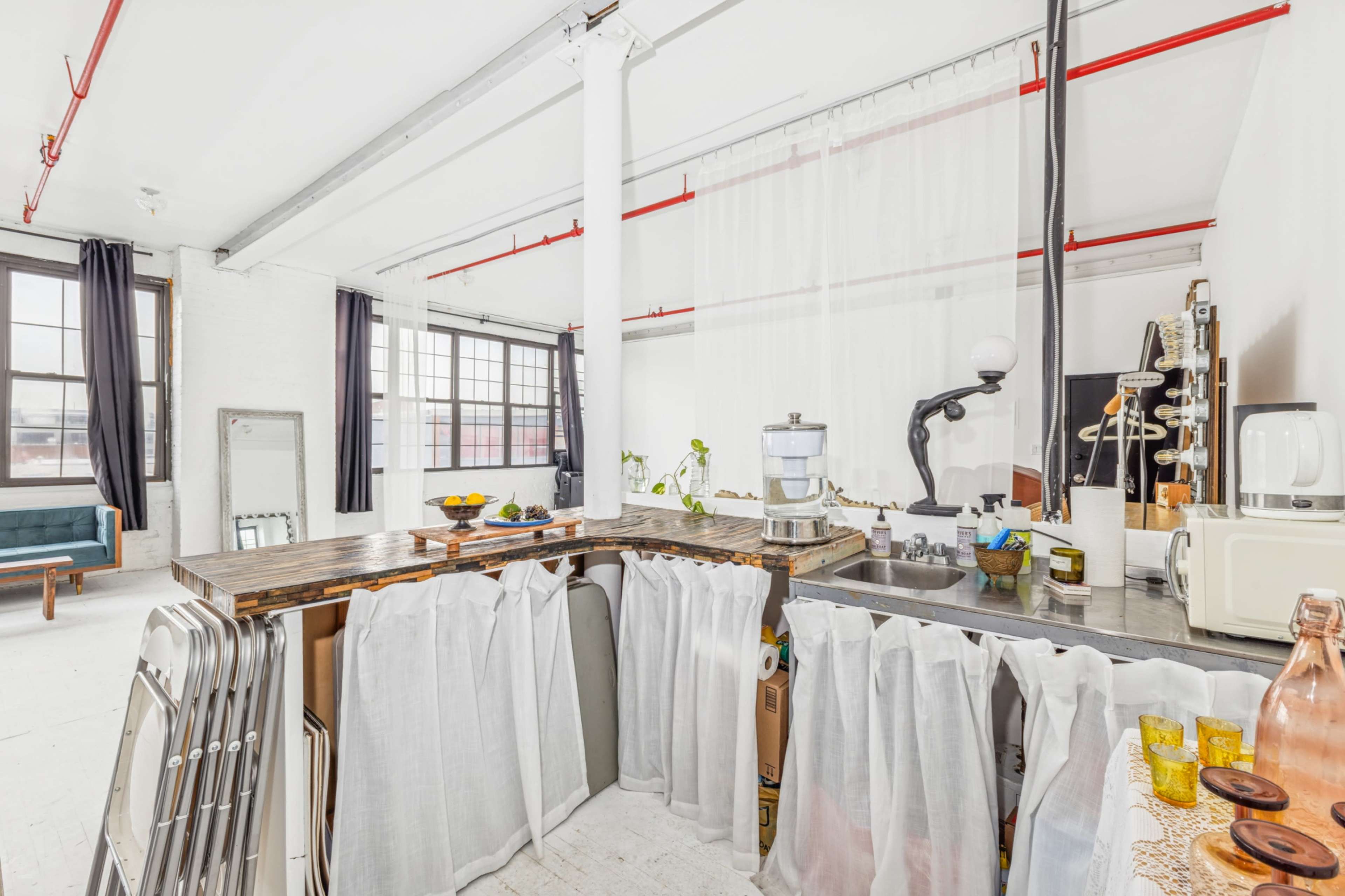 The image shows a bright, open kitchen area with a sink, a counter covered by white curtains, and large windows allowing natural light into the space.