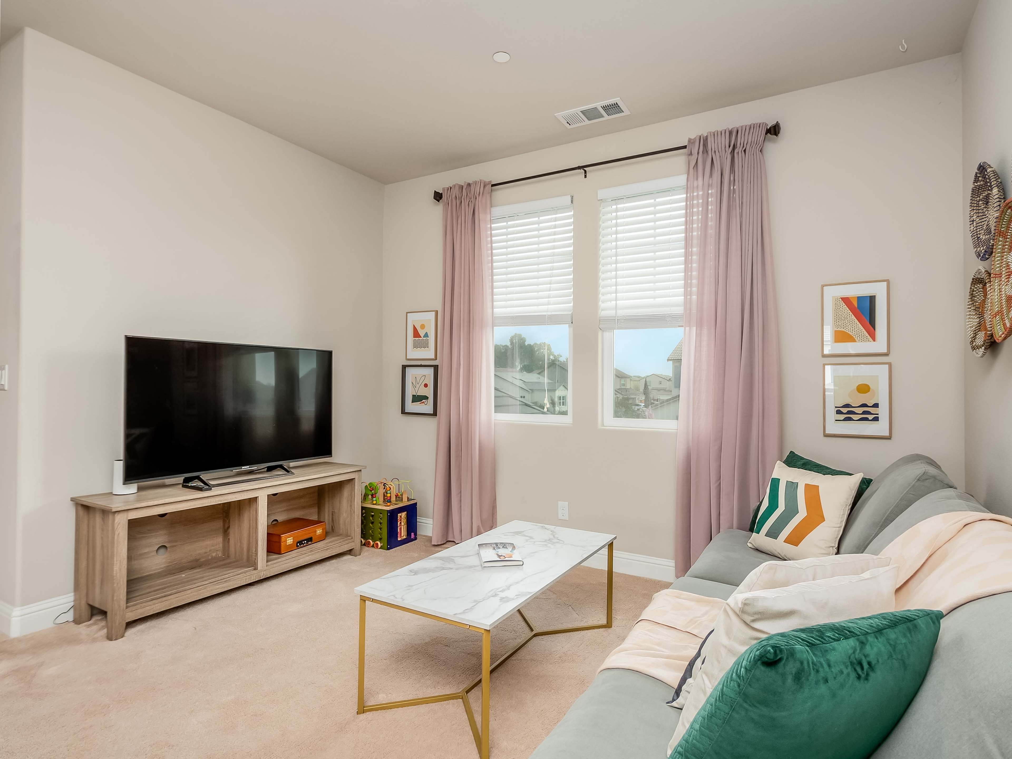 A living room with a green couch, a marble-top coffee table, a television on a wooden stand, and large windows covered with light curtains.