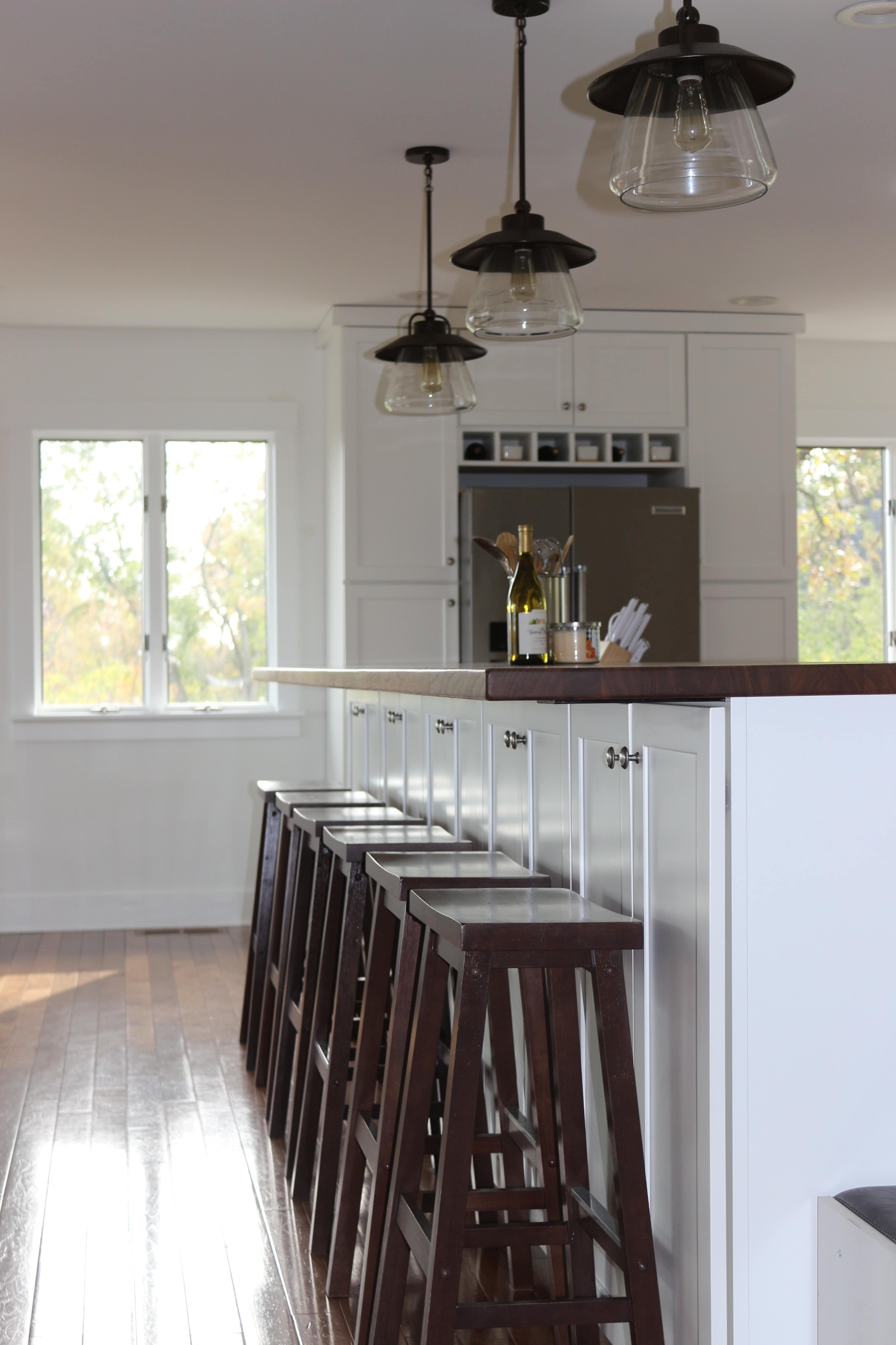 A modern kitchen with a white island bar and several wooden stools, beneath pendant lights, and large windows letting in natural light.