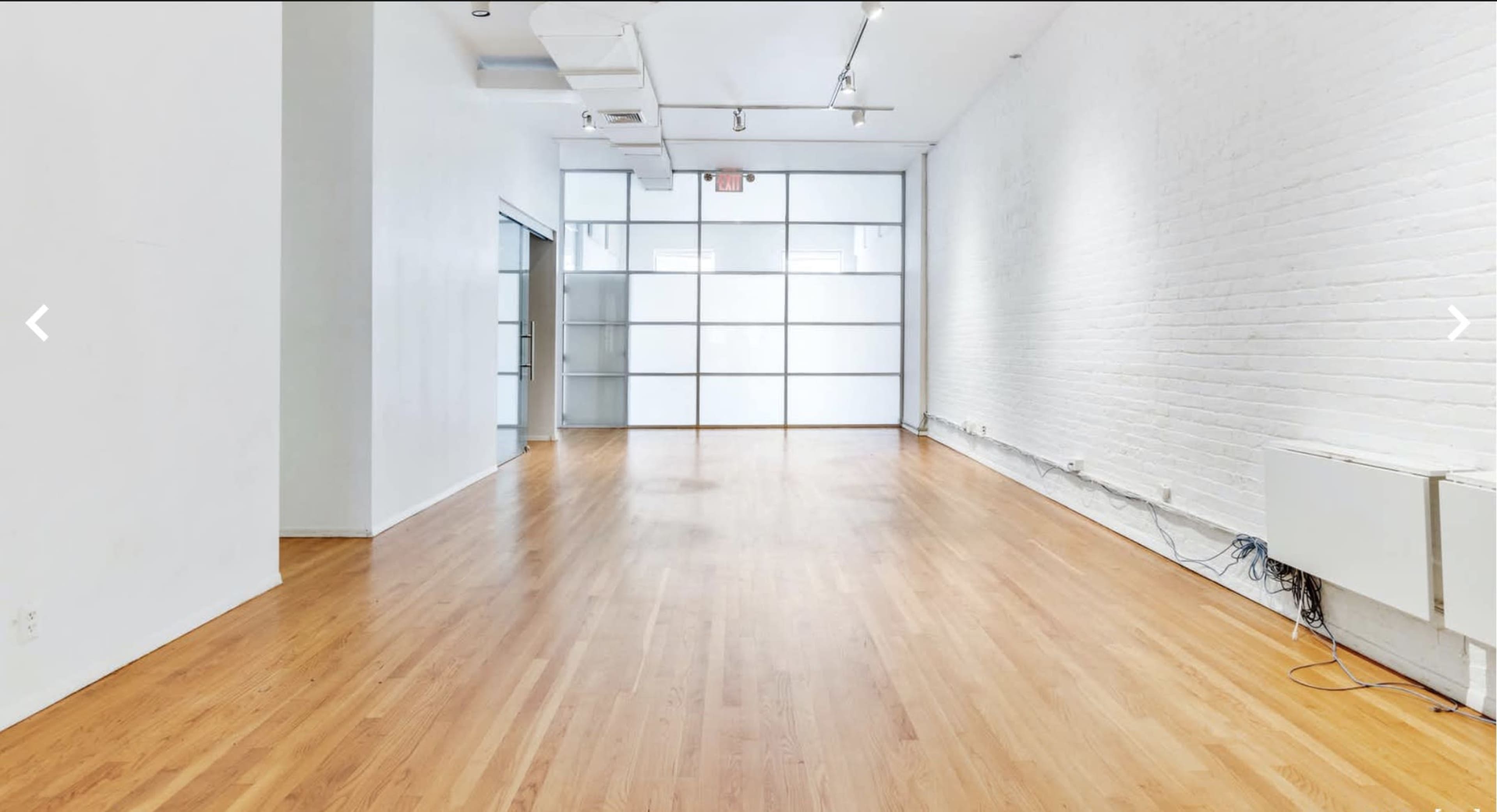A spacious, empty room with wooden flooring, white walls, and large windows at one end.