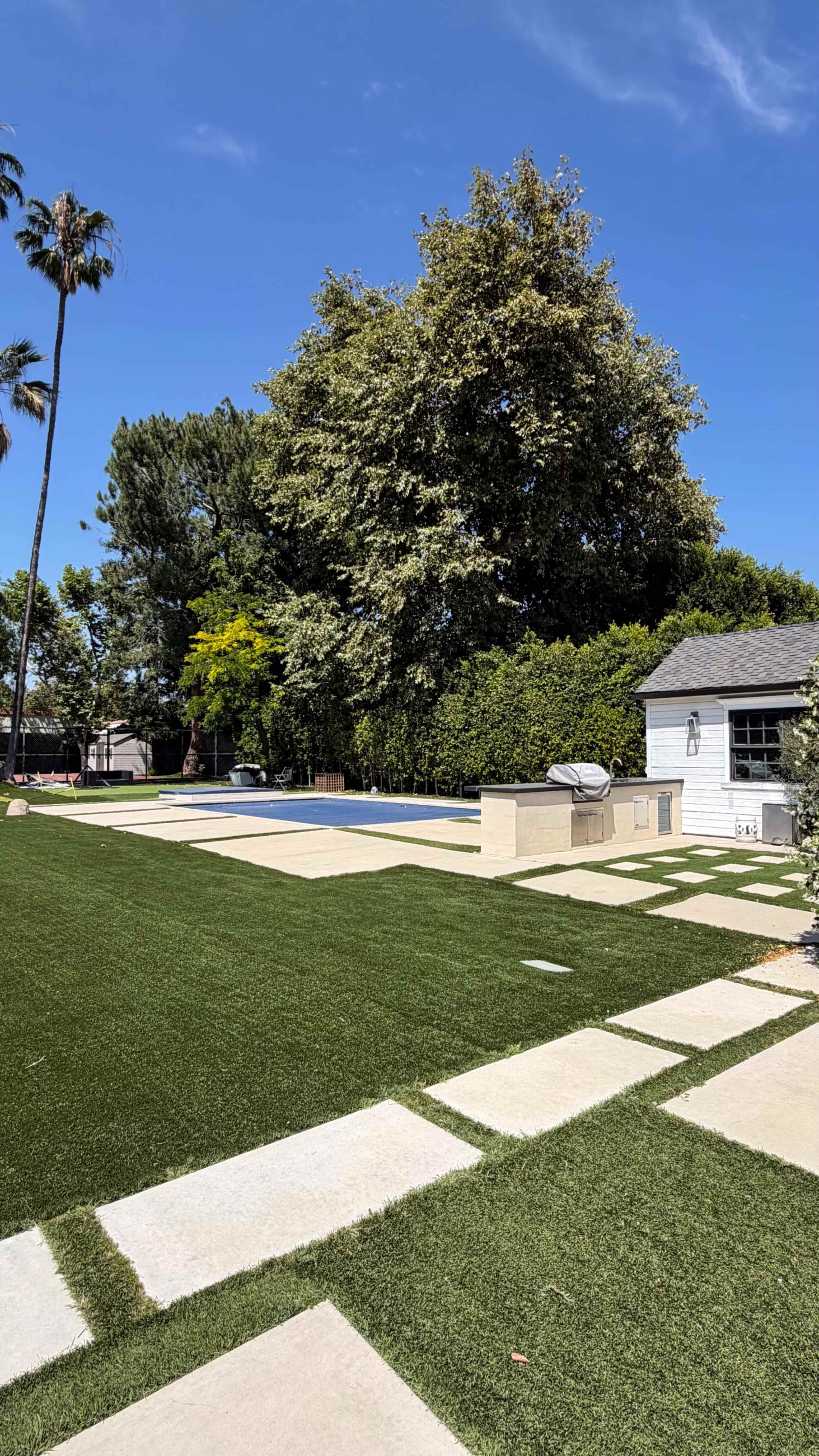 The image shows a backyard with a swimming pool, a patio area, and landscaped grass bordered by stone pathways.